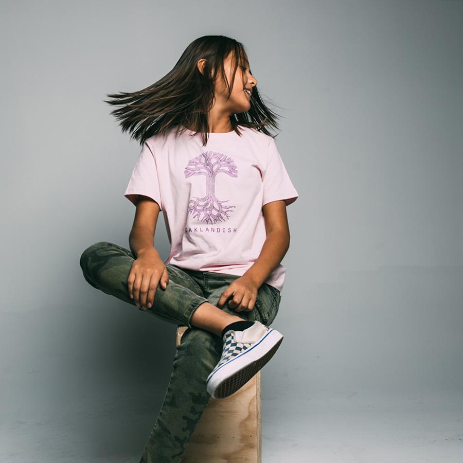 A young person with long brown hair, wearing a Youth Oaklandish Classic Logo Tee in light pink, featuring a purple tree design and the word "OAKLANDISH" below it, sits on a wooden box. They are smiling and looking to the side. They are also wearing camo pants and white sneakers with a blue pattern. The background is grey.