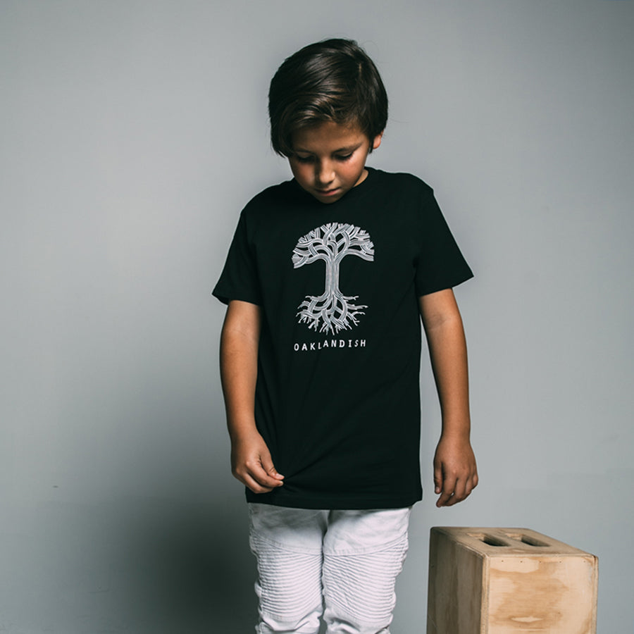 A young boy stands against a grey background, looking down and slightly to the left. He wears an Oaklandish Youth Classic Logo Tee made from 100% black cotton, featuring a white tree design with "OAKLANDISH" printed below. Clad in white pants, he stands next to a light wooden box.