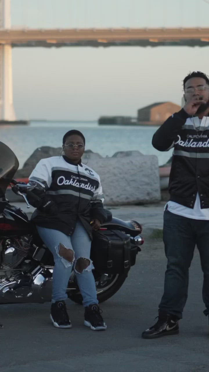 A man and a woman sitting outdoors in striped satin jackets with a California Oaklandish wordmark and Oaklandish tree logo.