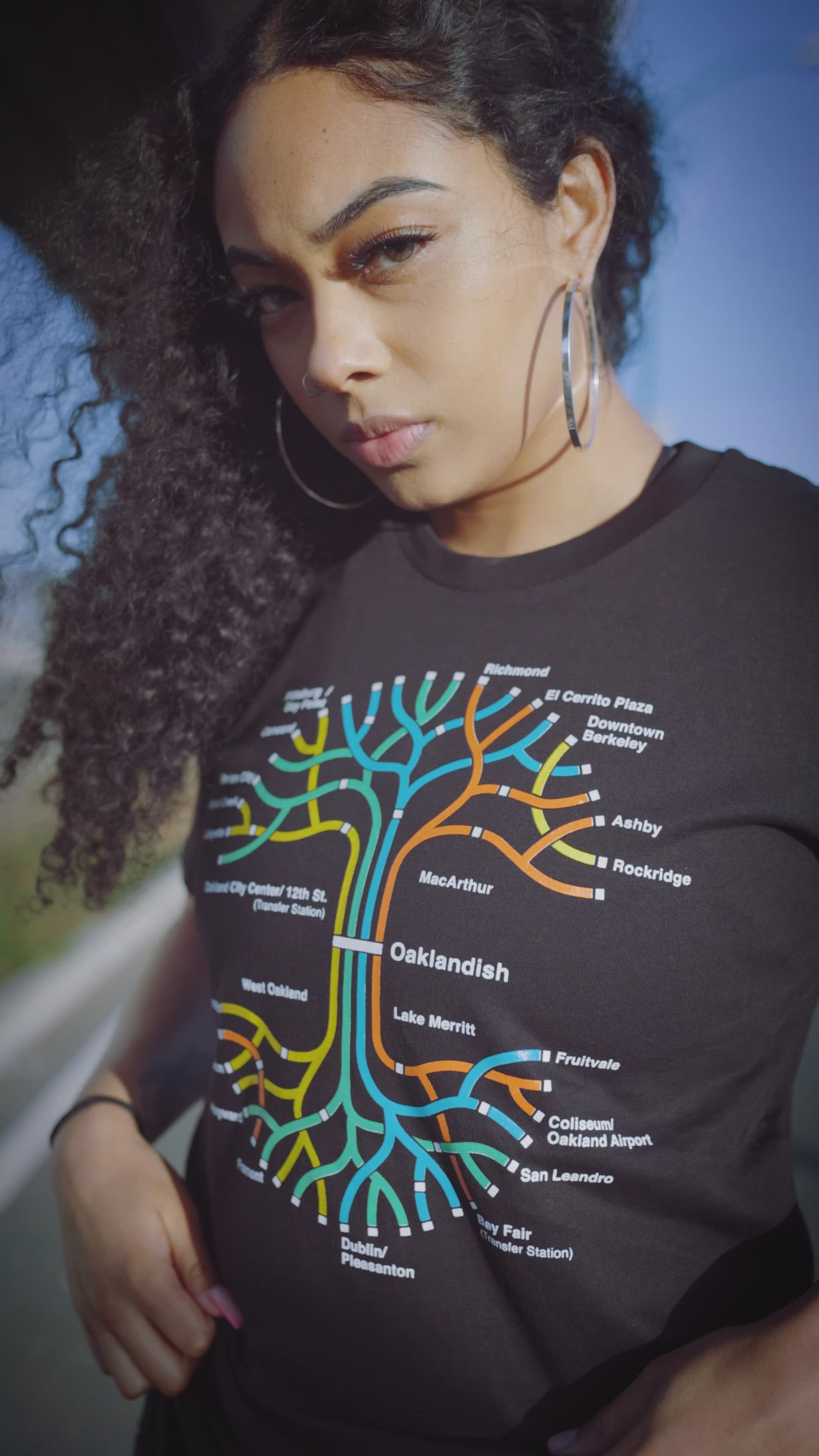 Woman standing under an overpass wearing a black t-shirt with a BART transit map in the shape of the Oaklandish tree logo.