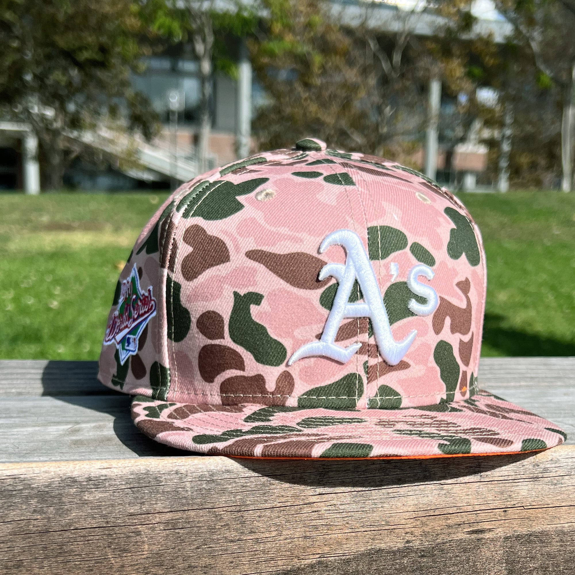 Duck camo cap with an embroidered Oakland As the logo on the crown and World Series 1989 patch on the right wear side sitting on an outdoor picnic table.