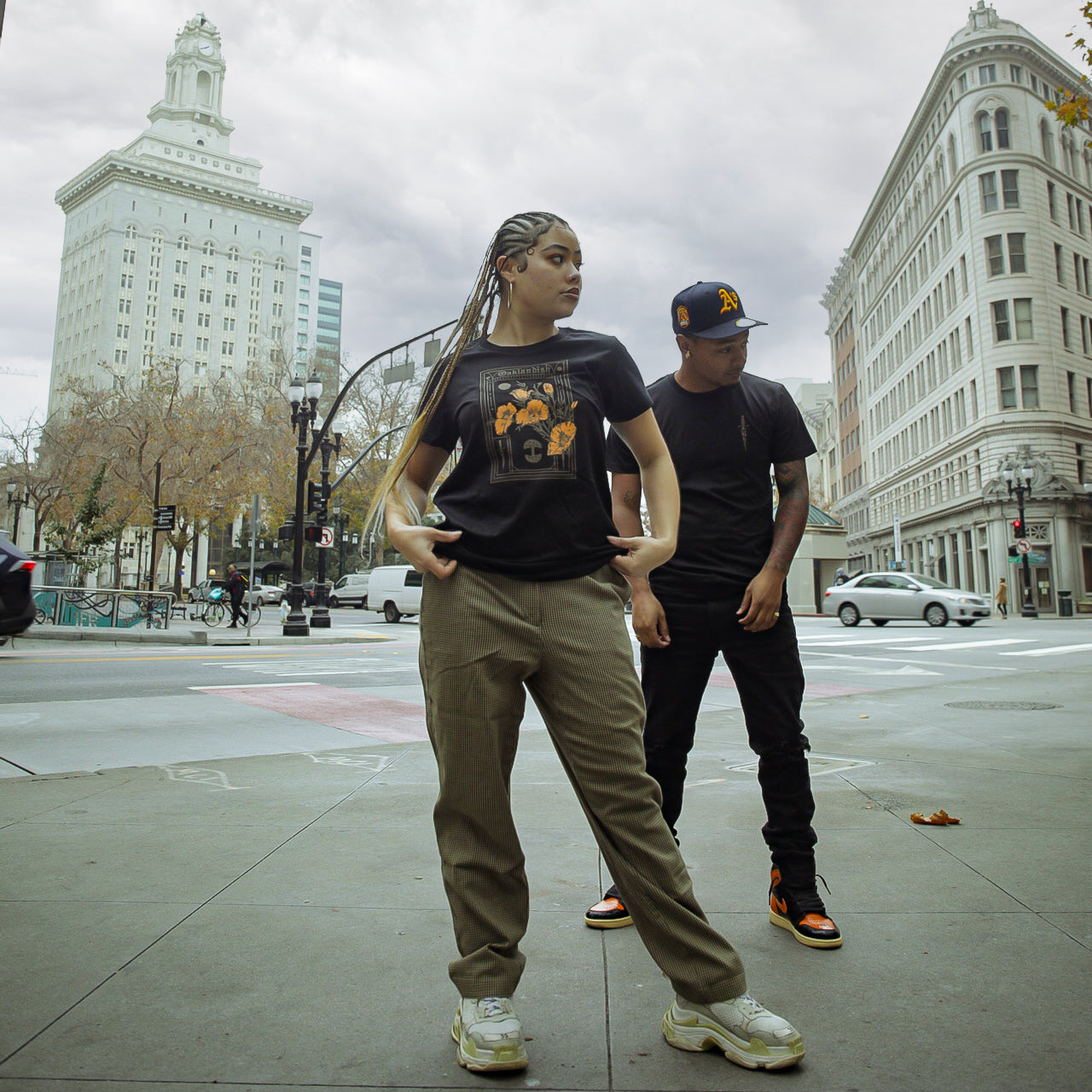 A man and woman standing on an Oakland street wearing black Oaklandish Blossom t-shirts.