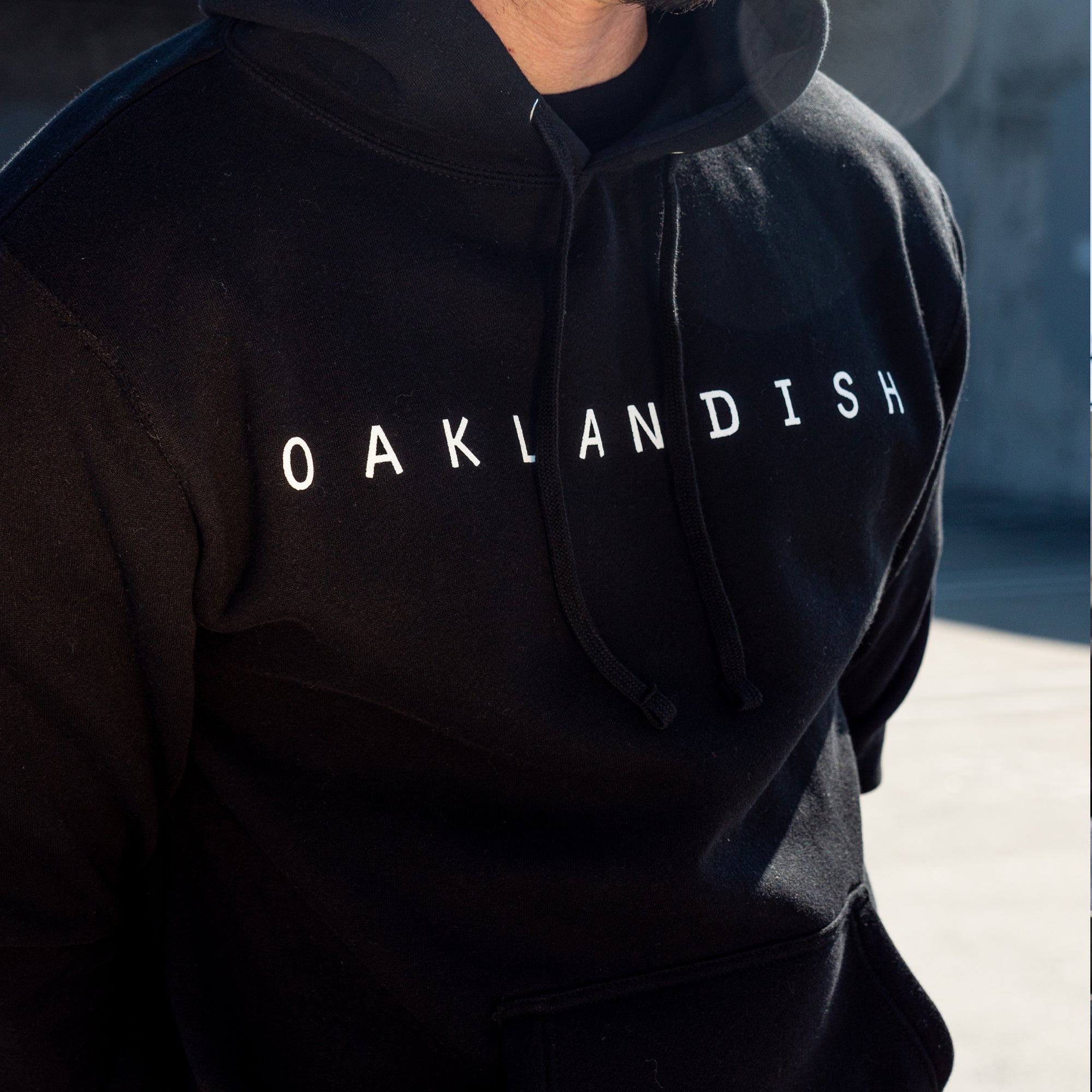 A person is partially visible from the neck to waist, wearing an Oaklandish Classic Hoodie in black with "OAKLANDISH" printed in white capital letters across the chest. The background is partially blurred and appears to be outdoors with sunlight casting shadows, highlighting their Oakland roots.