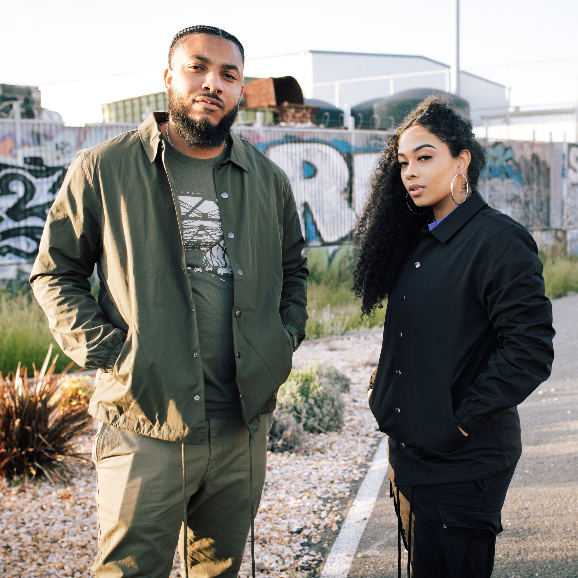 Man & woman in urban area with graffiti in background. Man is wearing green & woman is wearing a black cotton, collared, snap close jacket.