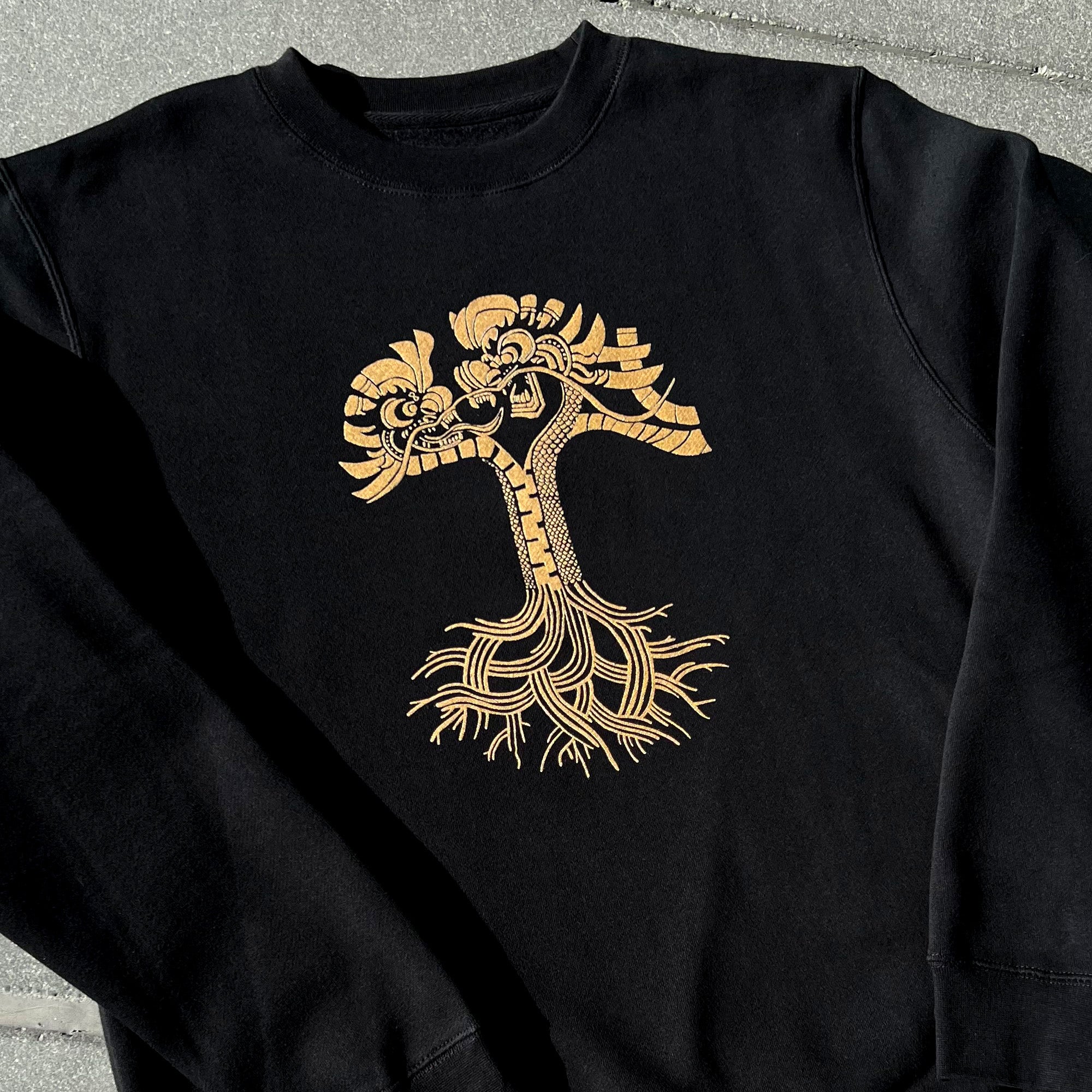 Black crewneck sweatshirt with metallic gold dragon power design in the shape of the Oaklandish tree logo outdoors on asphalt.