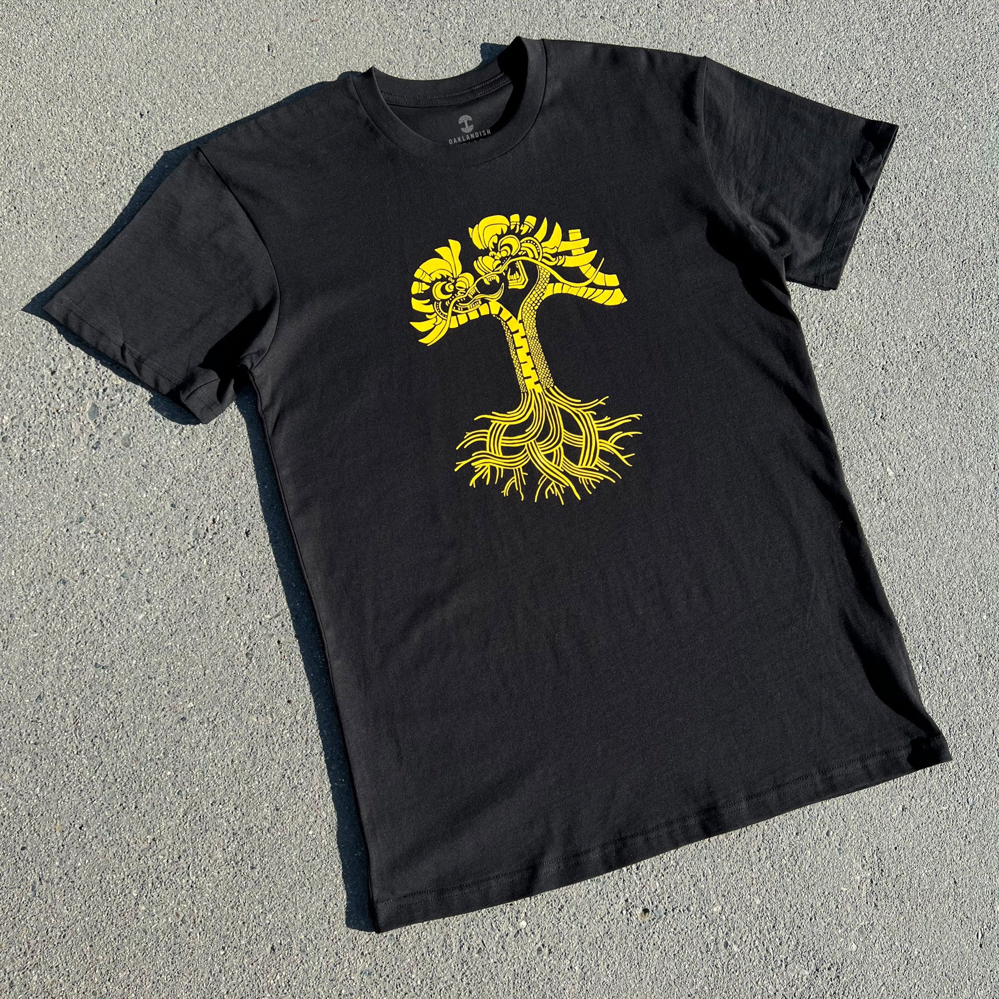 Black t-shirt with gold dragon power graphic design in the shape of an Oaklandish tree logo laying outdoors on asphalt.