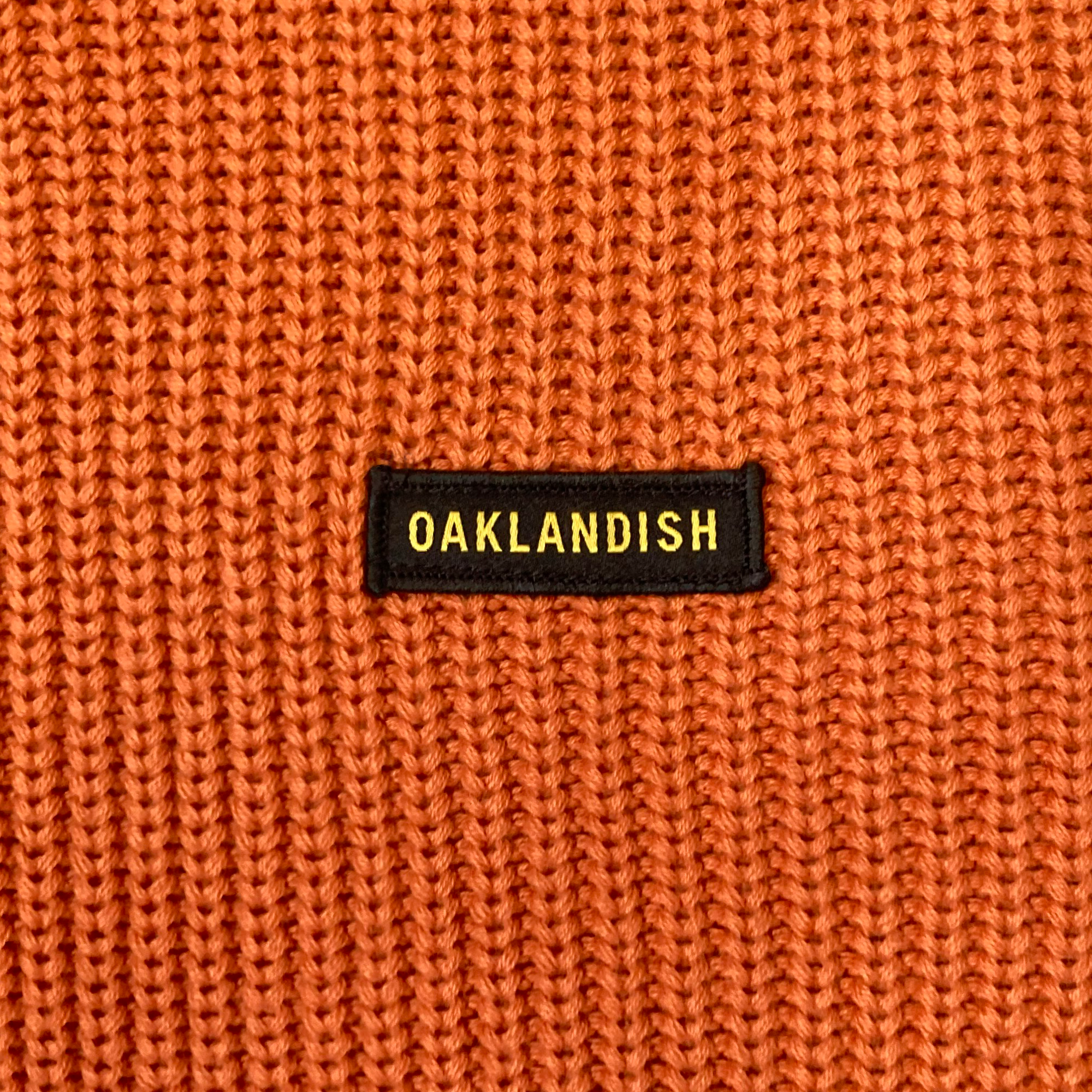Detailed close-up of embroidered Oaklandish wordmark woven label patch on an autumn orange heavy-knit sweater.