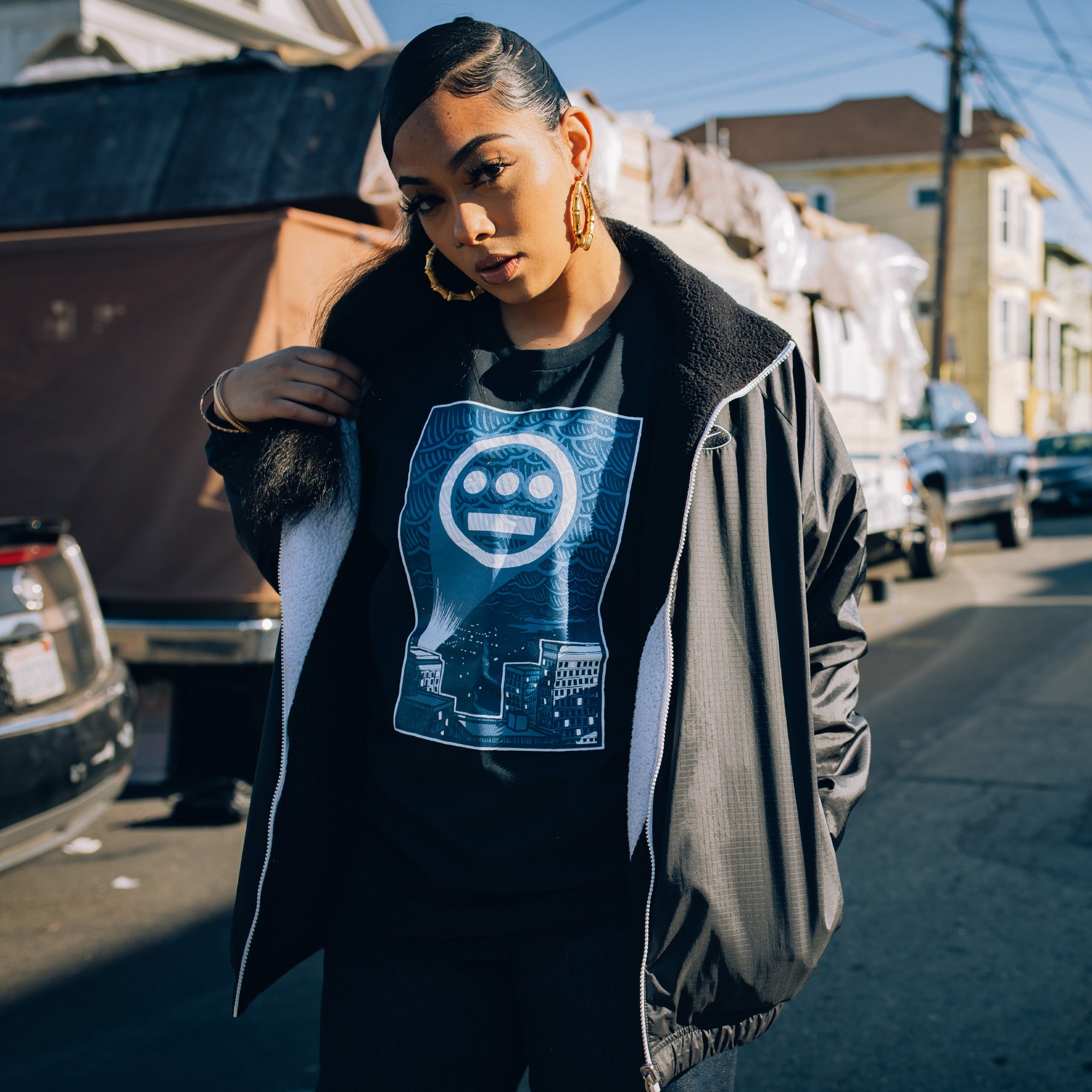 Female model wearing navy t-shirt with a graphic of Hiero logo spotlight signal in Oakland’s night sky above buildings.