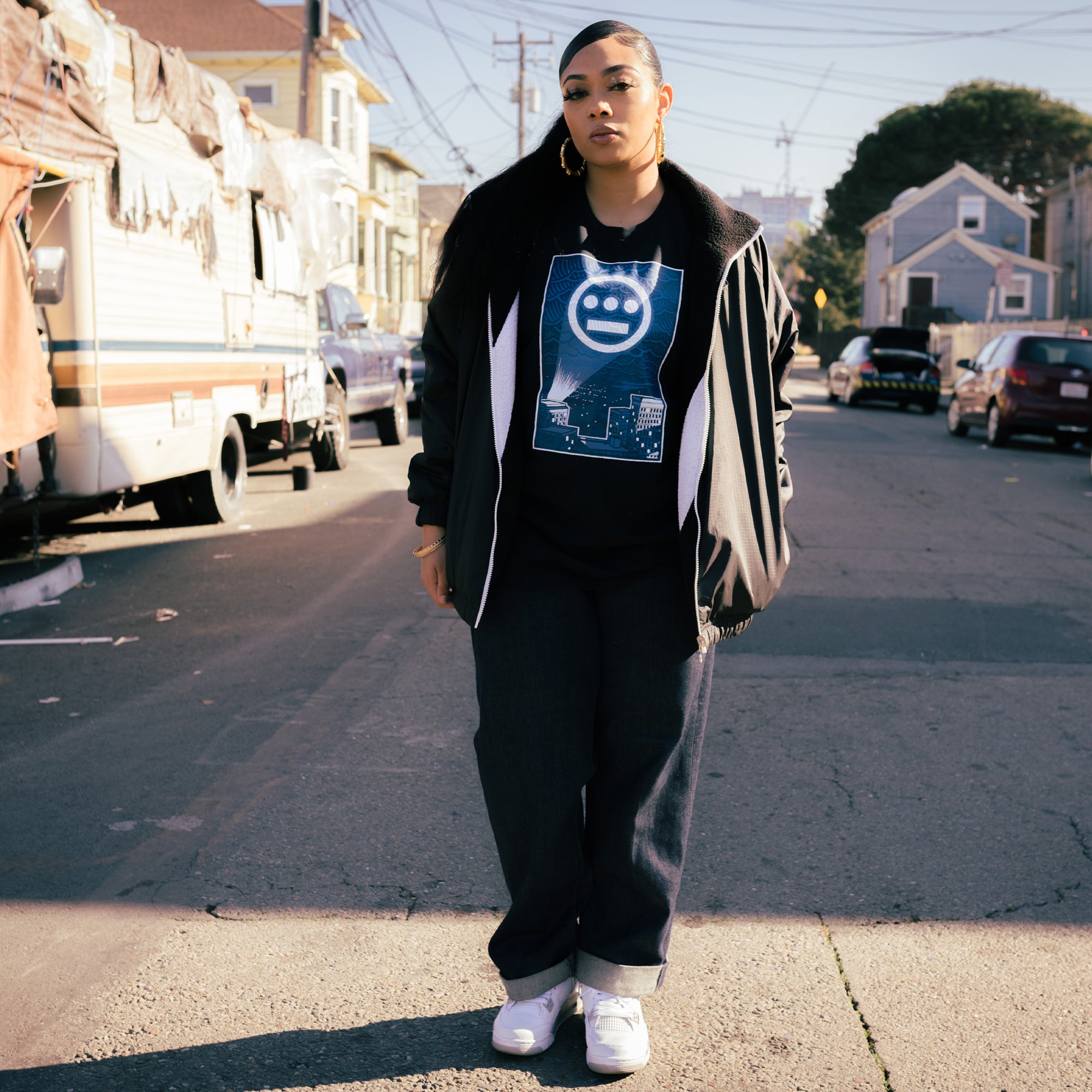 Female model in the middle of the street wearing a navy t-shirt with a graphic of Hiero logo spotlight signal in Oakland’s night sky above buildings.