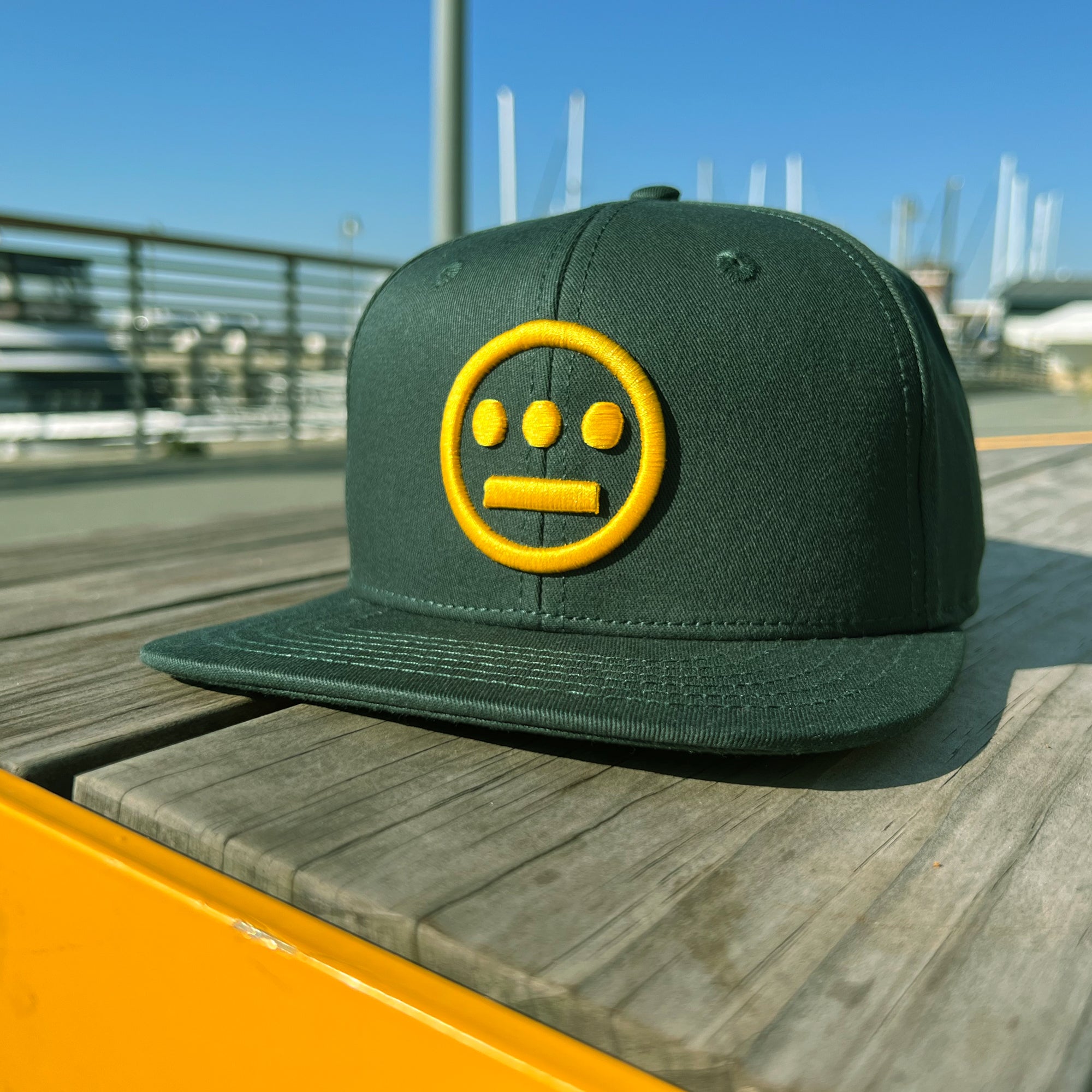 The Hiero Logo Snapback by Hieroglyphics, in dark green, rests on a wooden surface outdoors. The front features a yellow embroidered logo resembling hieroglyphics with a horizontal line and circles above and below it. In the background, a marina with boats and a blue sky is visible.