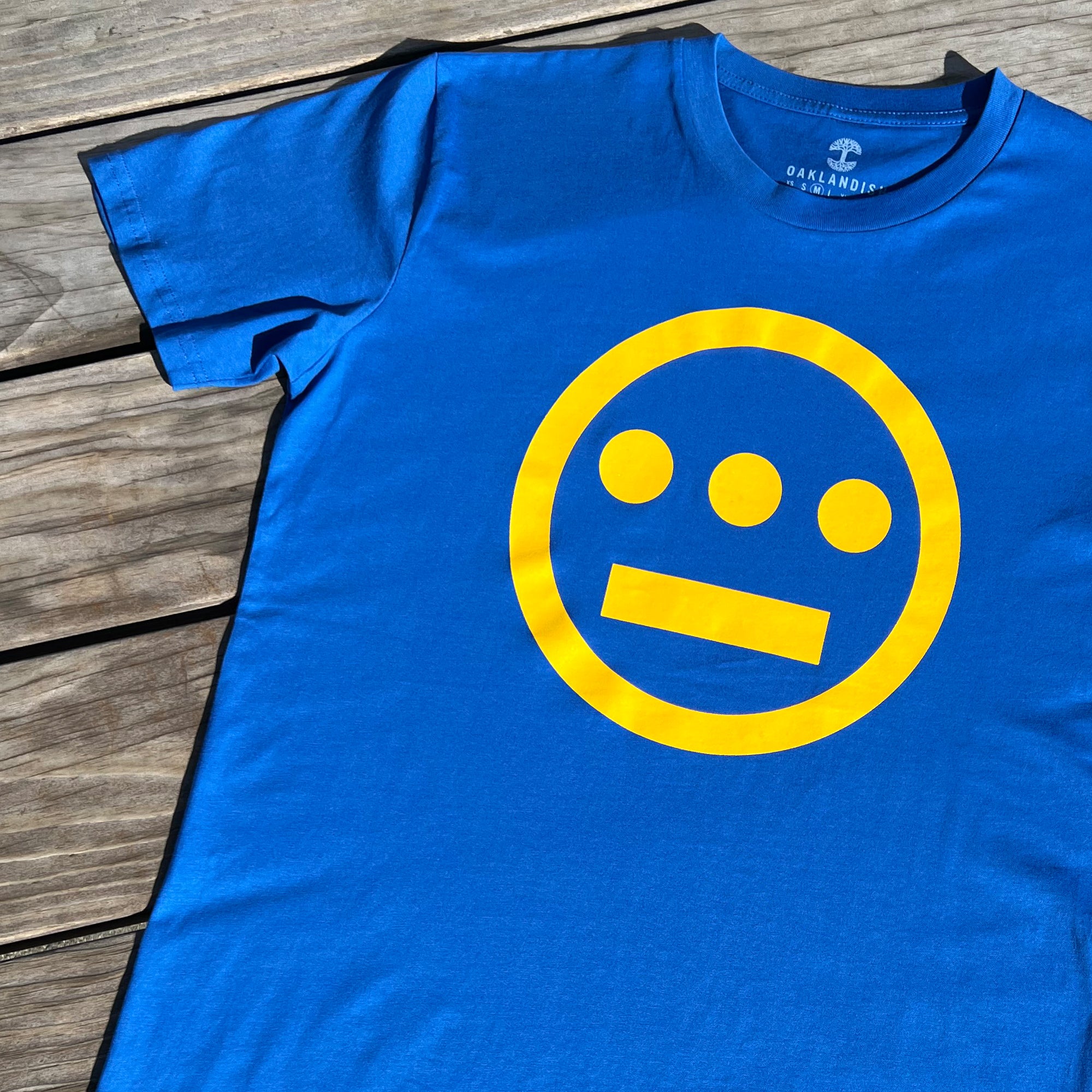 Close up of yellow Hieroglyphics Hip-Hop logo on a royal blue t-shirt on a wooden deck.