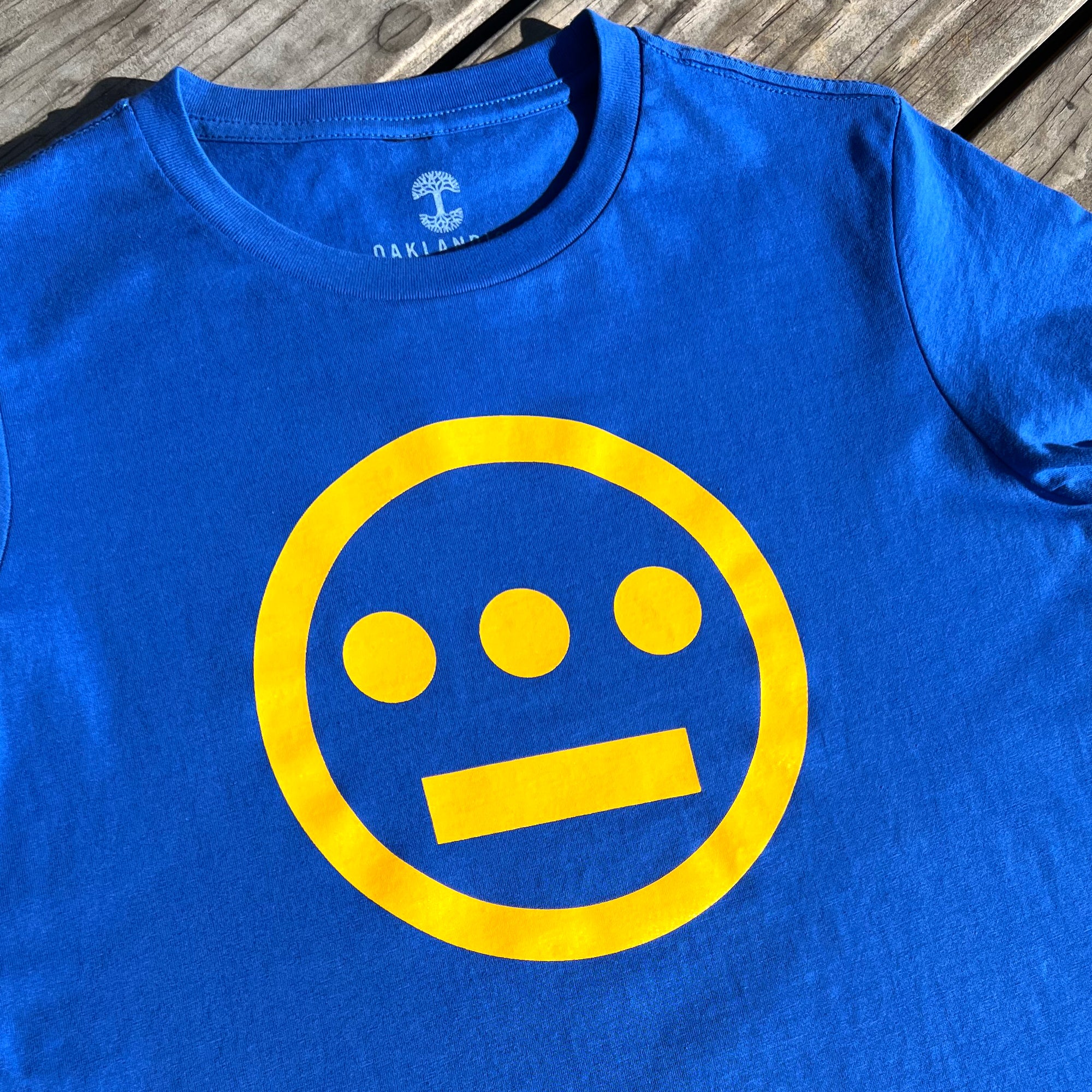 Close up of yellow Hieroglyphics Hip-Hop logo on a royal blue t-shirt on picnic bench.