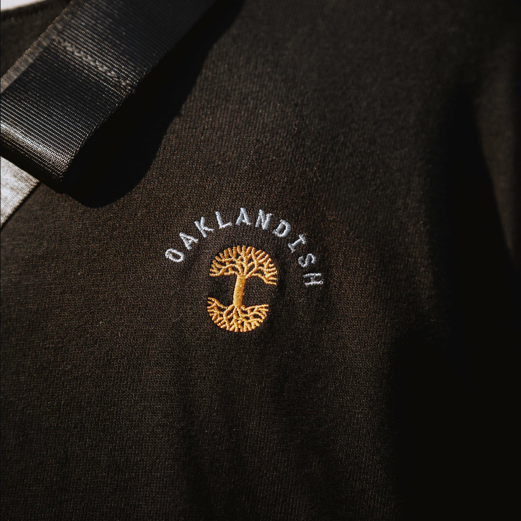 Close-up on model  of embroidered gold Oaklandish tree logo, white wordmark on the chest of a black long-sleeve t-shirt.