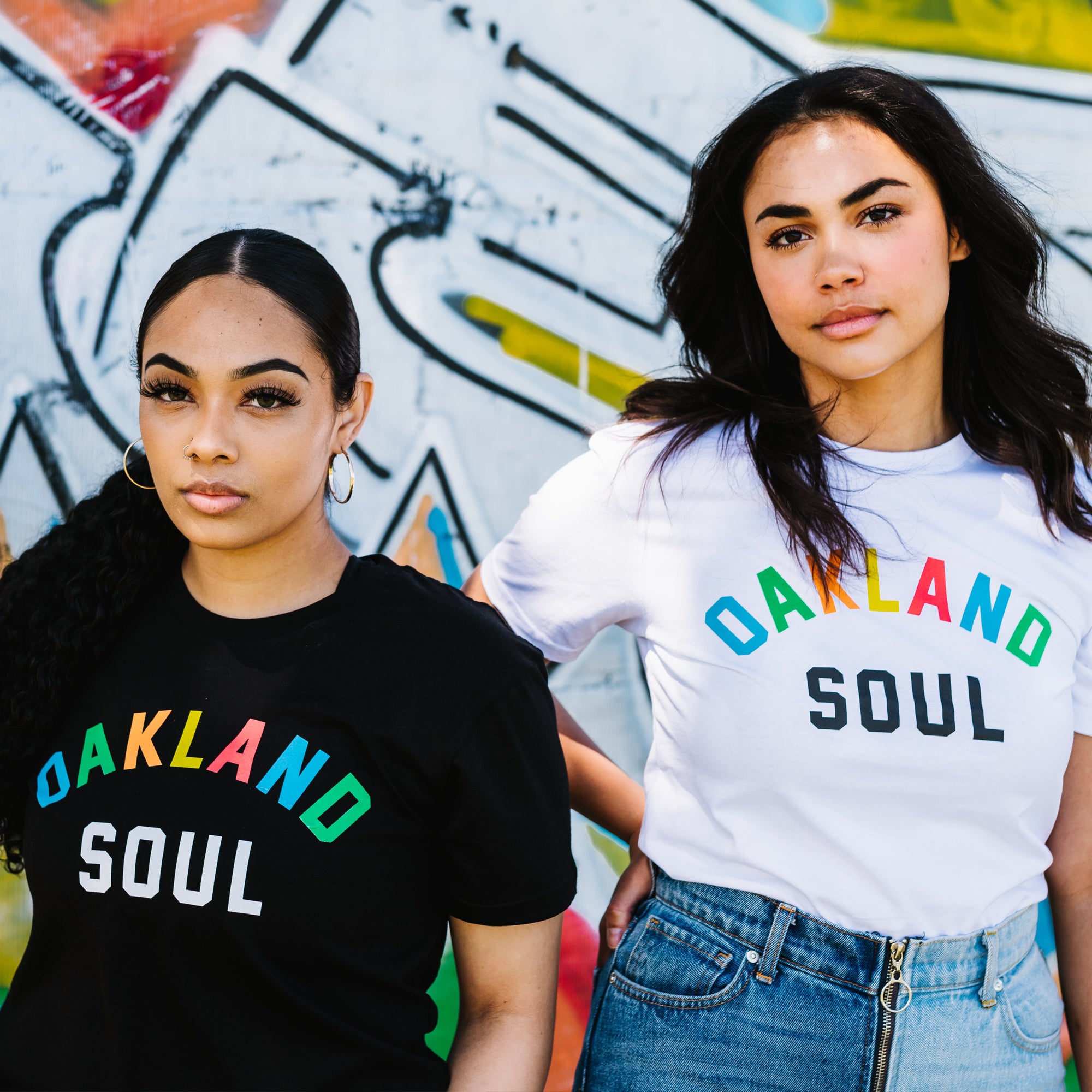 Two woman standing with waist up view & graffiti background. Both wearing Oakland Soul Wordmark T-Shirts.