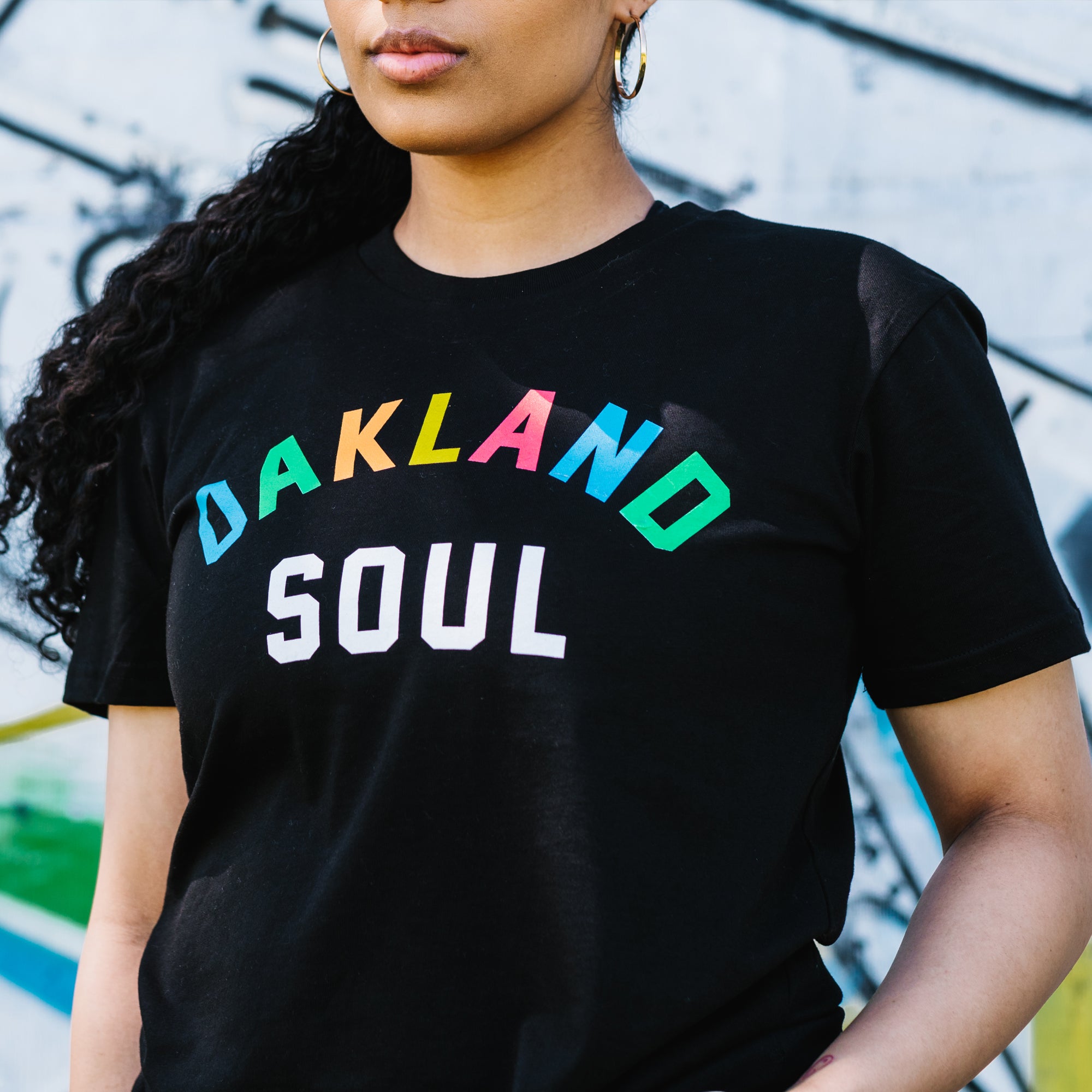 Close up of woman from waist to chin, wearing black t-shirt with full color Oakland Soul wordmark logo. Blurred graffiti background.