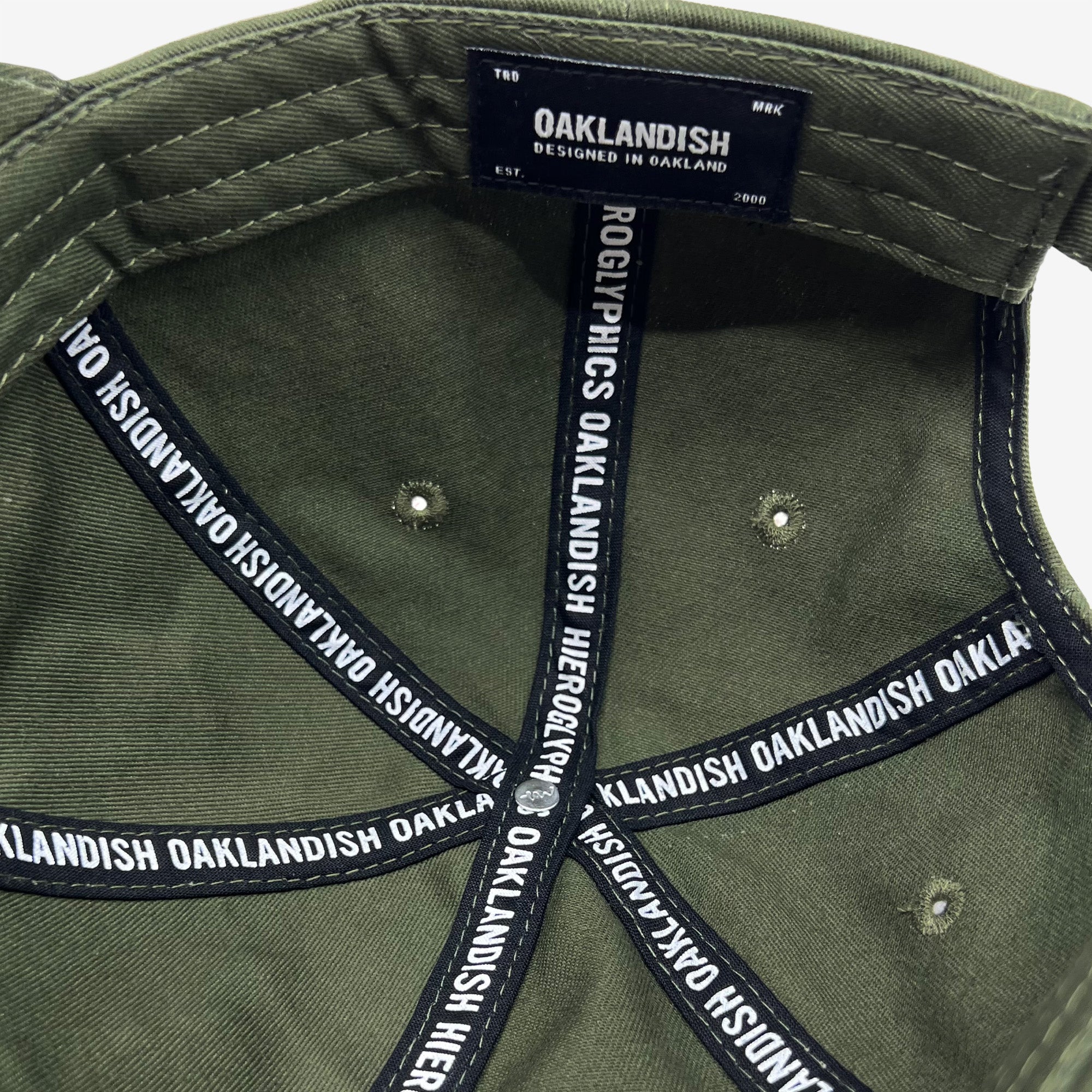 Close-up view of the interior of an olive green Official Micro Logo Dad Hat. The black sweatband inside features white text reading "OAKLANDISH" and "HIEROGLYPHICS." A black label with similar branding and "DESIGNED IN OAKLAND" is sewn into the inner back above the adjustable strap.