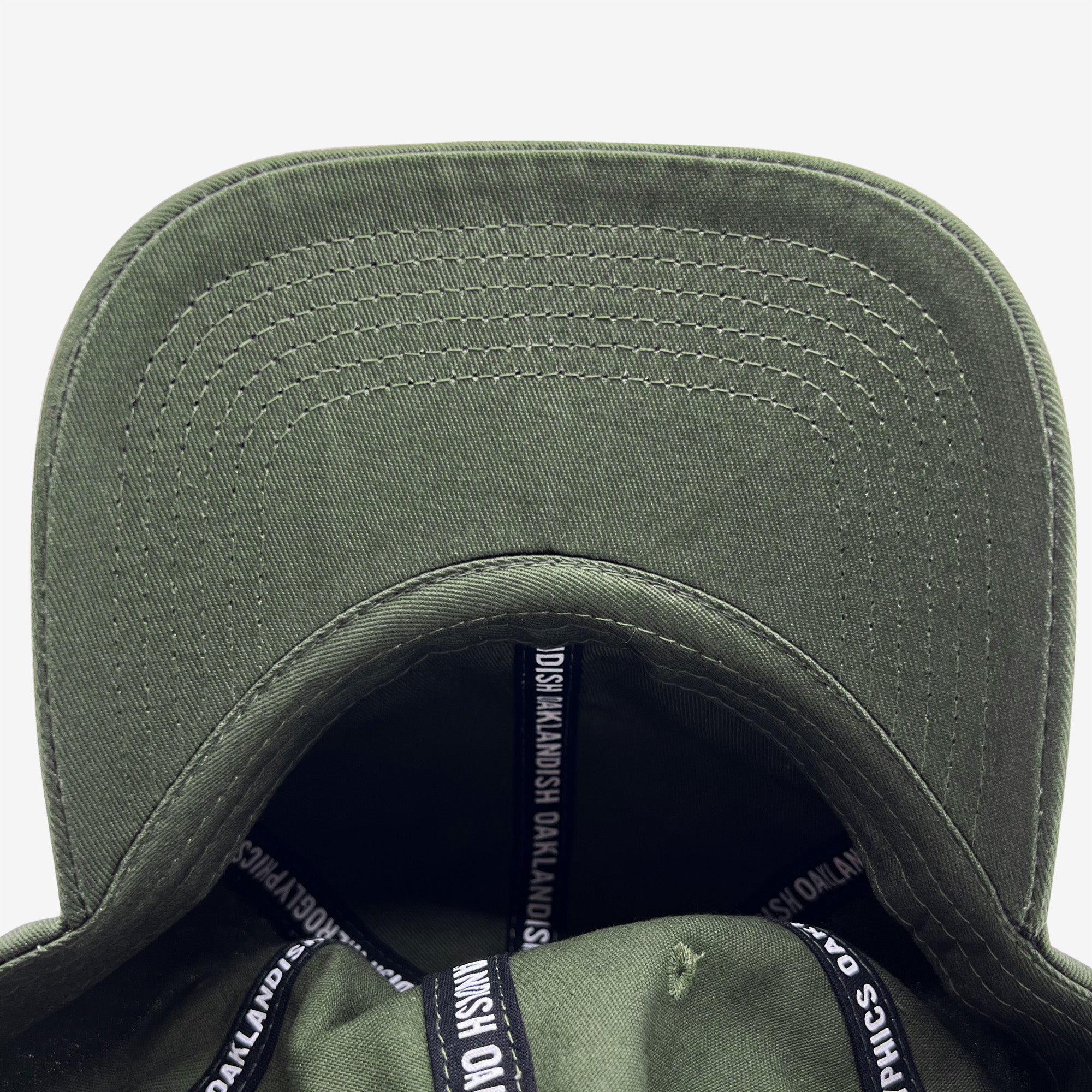 A close-up image of the underside of the Official Micro Logo Dad Hat by Oaklandish in army green. The inside features black tape with white text that reads "OAKLANDISH." The stitching on the green fabric is visible, showcasing a clean and structured design, while an adjustable strap ensures a comfortable fit.