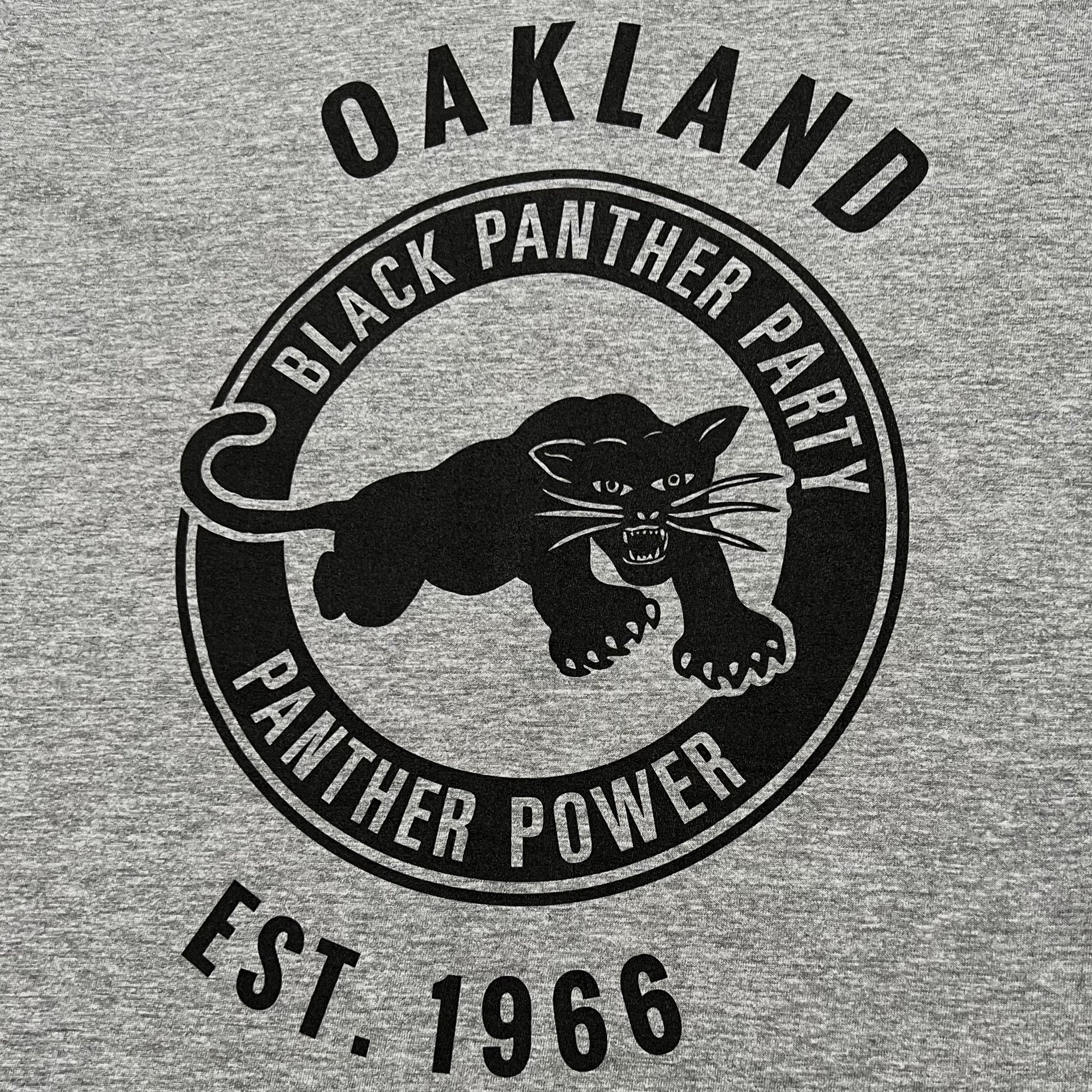 The Black Panther Power Tee by Oaklandish features a black design on a gray t-shirt with a central image of a panther in a pouncing position. The text "BLACK PANTHER PARTY" encircles the top of the panther, while "PANTHER POWER" is at the bottom. Above and below this circular design, additional text celebrates the influential community organization with "OAKLAND" and "EST. 1966.