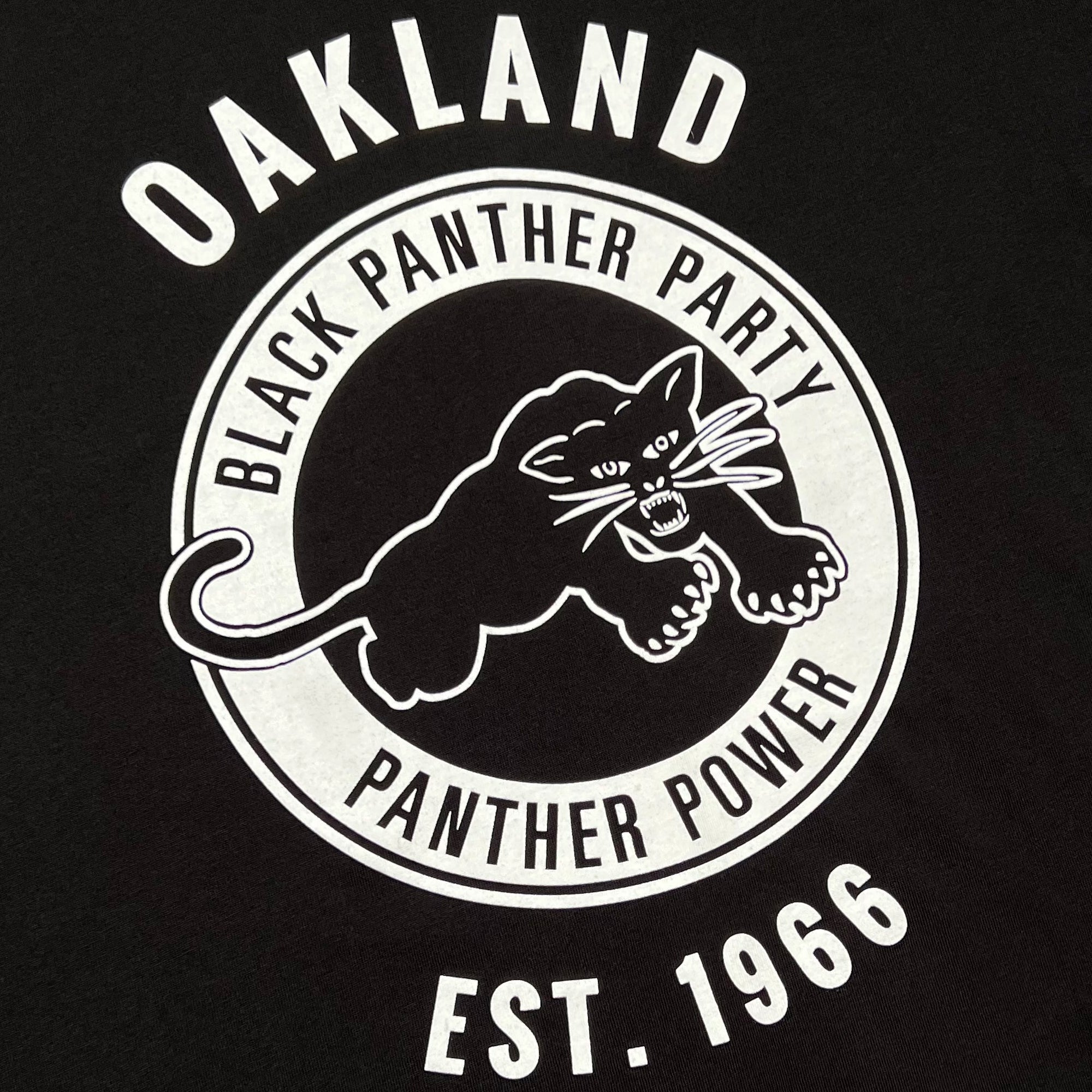 The **Black Panther Power Tee** by **Oaklandish** features a black background with a white circular insignia depicting a black panther illustration. The text around the insignia reads "Oakland," "Black Panther Party," "Panther Power," and "Est. 1966." The panther is shown in a pouncing position with bared teeth and extended claws, embodying Social Consciousness Apparel.