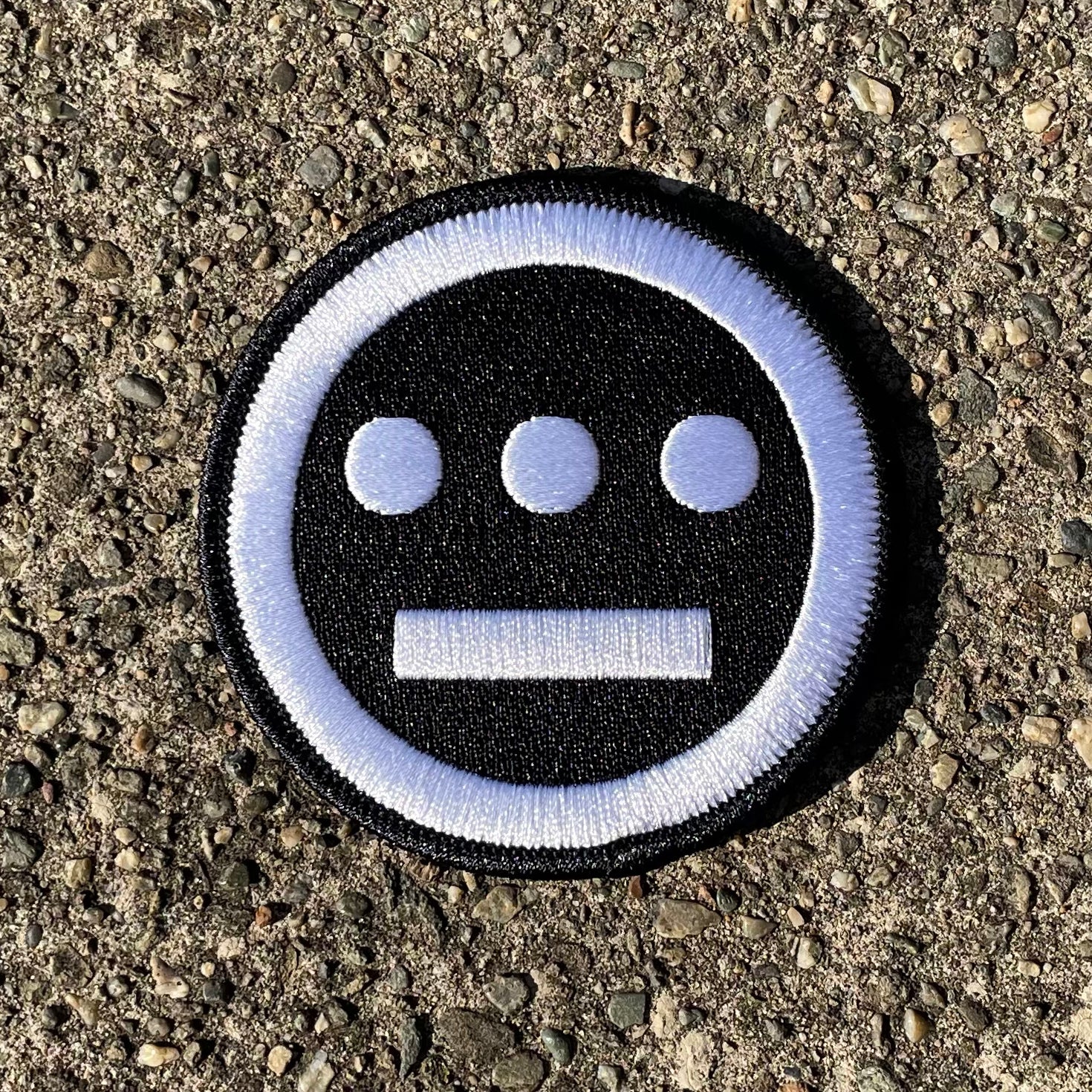 The Hiero Logo Patch by Hieroglyphics is a circular embroidered iron-on patch that features three white dots aligned horizontally above a horizontal white line on a black background, bordered by a white ring. Resting on rough, gray asphalt, its simple and bold design evokes modern hieroglyphics, making it a striking accessory.