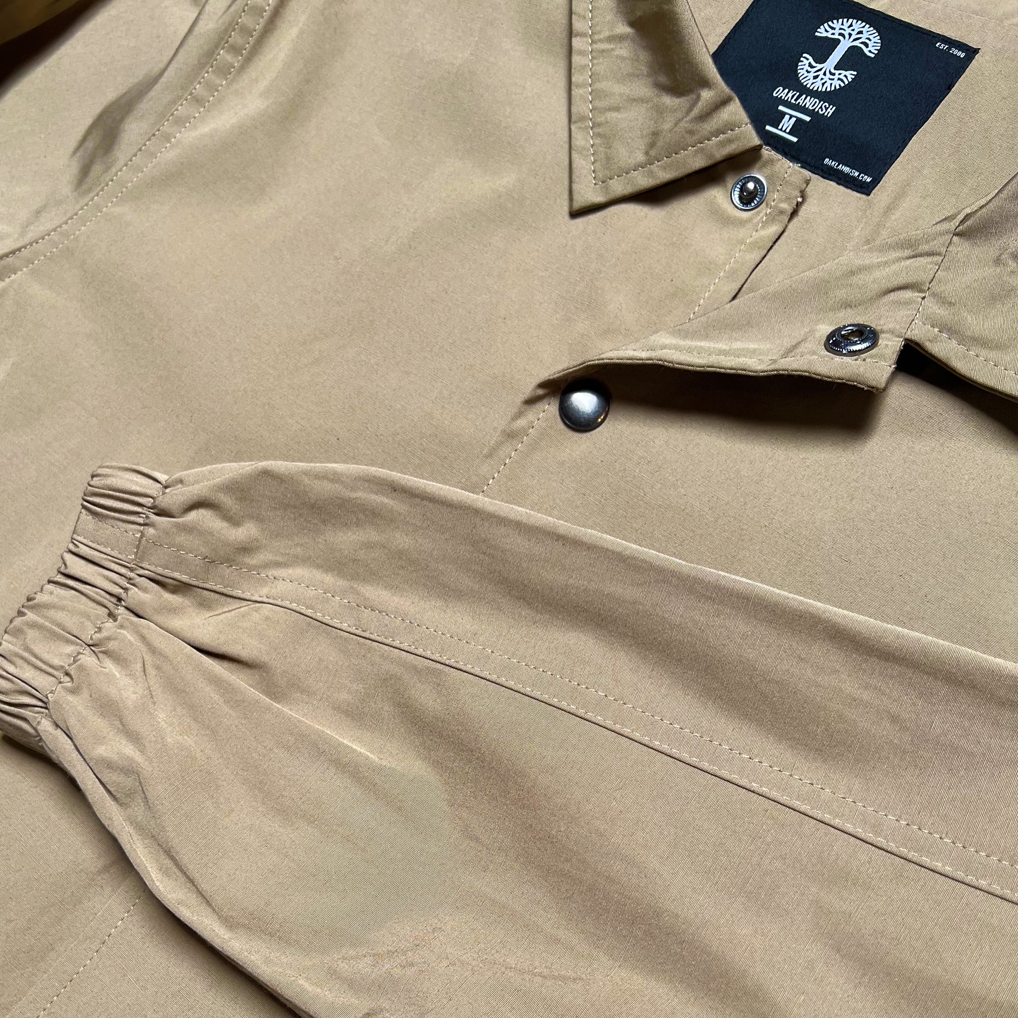 Close-up of the Premium Coaches Jacket from Oaklandish, a beige garment featuring a collared neckline with metal snap buttons and an adjustable wrist cuff with elastic. Inside the neck, a black embroidered tag displays a tree symbol and text. The fabric has a smooth, matte texture, indicative of its water-resistant nature as part of this men's casual or utility jacket.