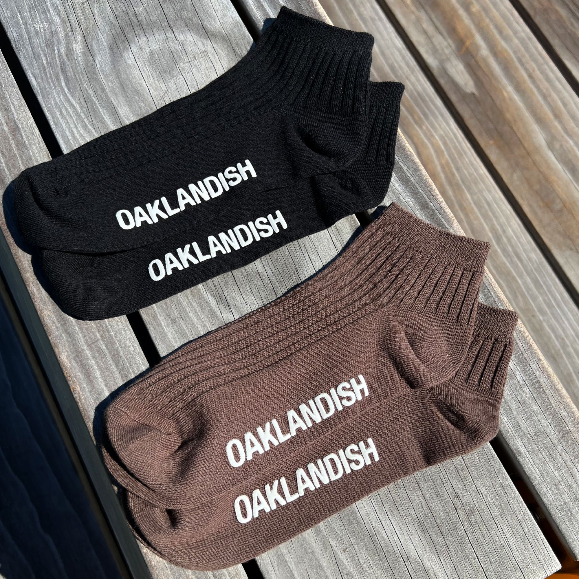 Two pairs of the Grip Low Crew Socks from Oaklandish on a wooden surface. The top pair is black with "OAKLANDISH" printed in white, while the bottom pair is brown with "OAKLANDISH" printed in white. Both pairs have a ribbed texture. The natural, rustic appearance of the wood enhances the charm of these low crew socks.