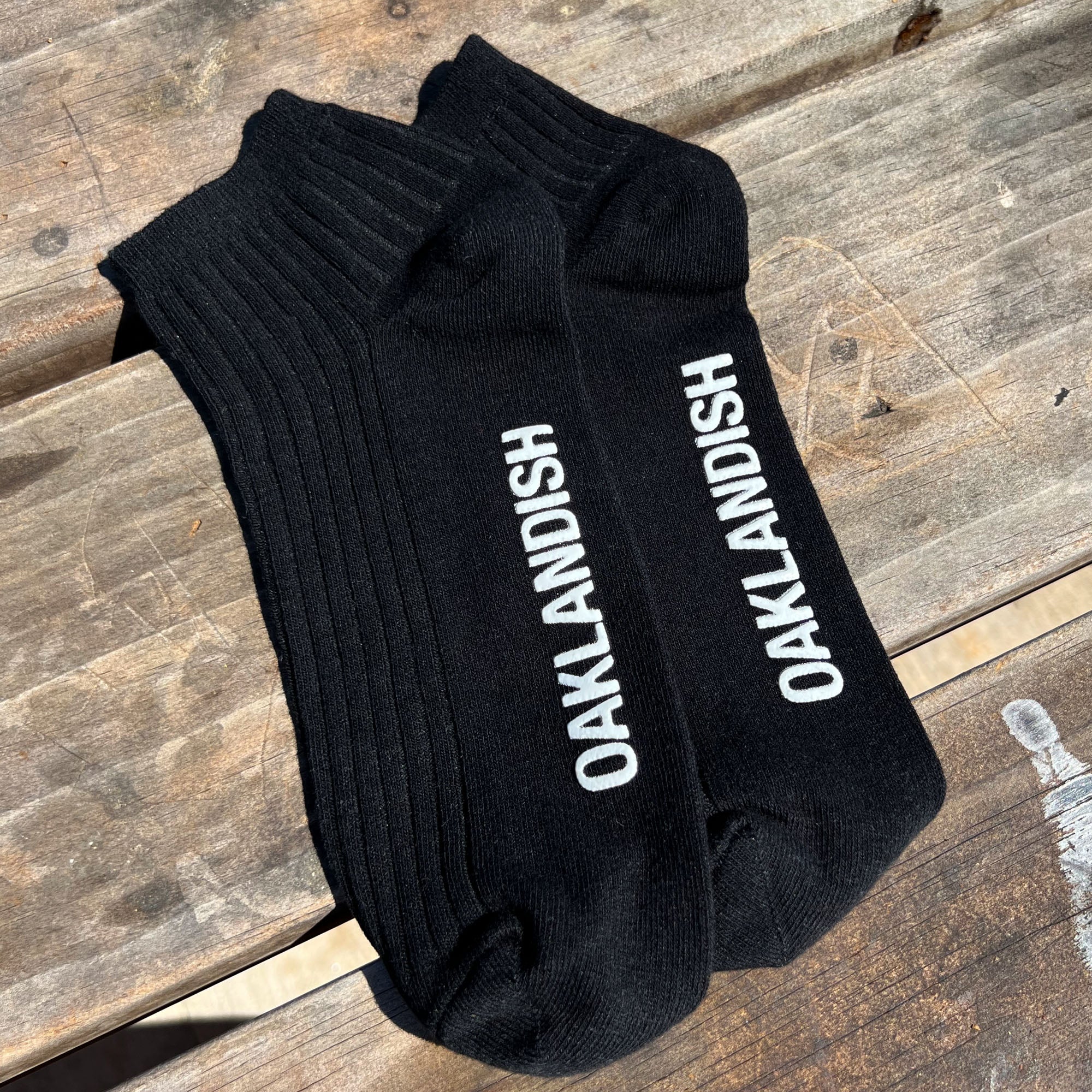 A pair of Oaklandish Grip Low Crew Socks, featuring the word "OAKLANDISH" written in white on the top of each black sock, is laid side by side on a rustic wooden surface. Sunlight casts shadows, accentuating the texture of the wood.