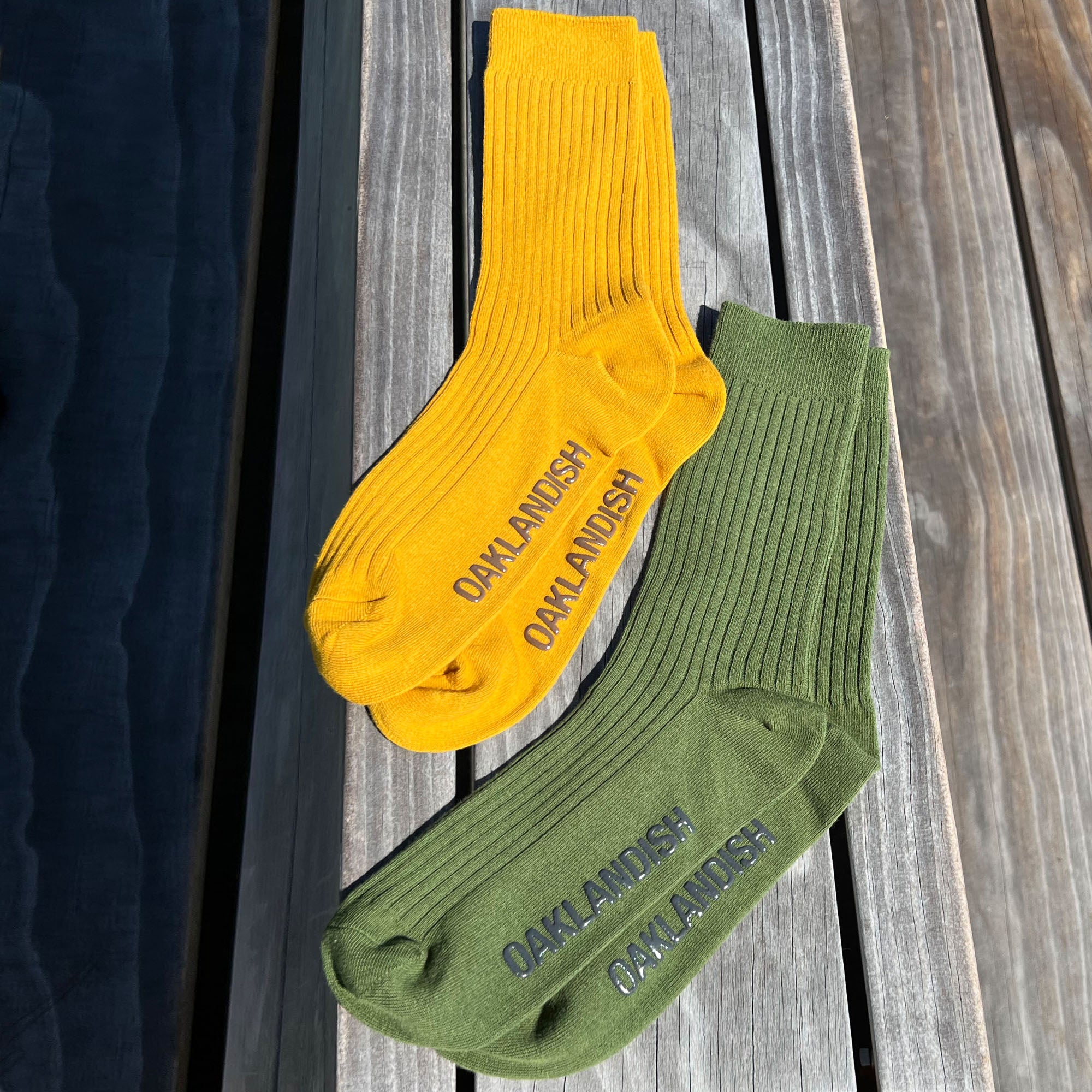 Two pairs of Oaklandish Grip Crew Socks are laid out on a wooden surface in the sunlight. One pair is bright yellow and the other is olive green. Made from a blend of cotton, polyester, and spandex, both pairs have "OAKLANDISH" printed in dark letters on the top side of the foot area.