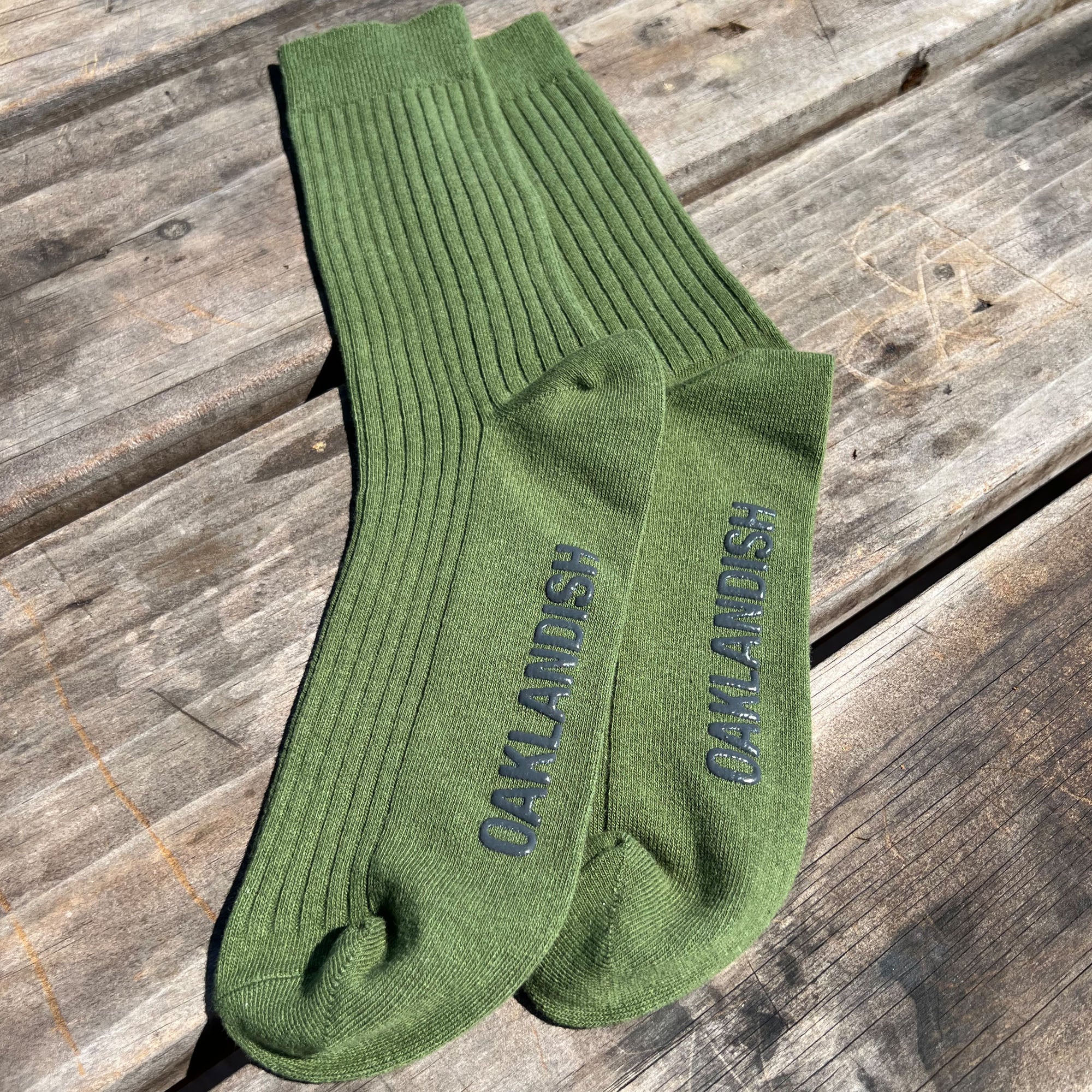 A pair of Oaklandish Grip Crew Socks in green, featuring ribbed grips and the brand name "OAKLANDISH" in dark letters, lie flat on a weathered wooden surface. The socks are positioned parallel with the brand name visible at the arch area of each sock. Sunlight casts shadows, emphasizing the texture of both the socks and the wood.