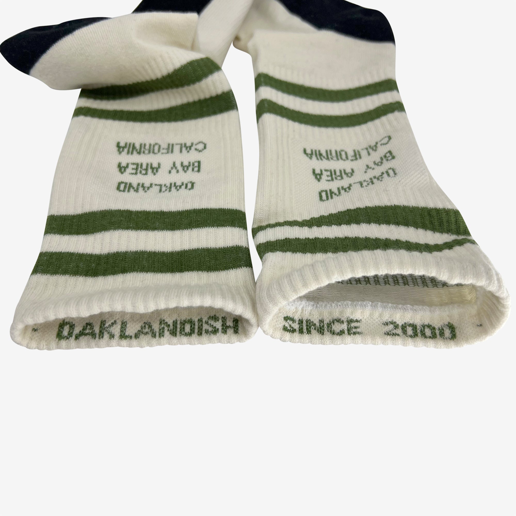 A close-up image of a pair of Bay Area Stripes Socks by Oaklandish, featuring green stripes and the text "OAKLANDISH, BAY AREA, CALIFORNIA" printed on the sides. Near the toes, "OAKLANDISH" and "SINCE 2000" are also printed. These comfortable cotton socks for adults are slightly folded and positioned side by side.