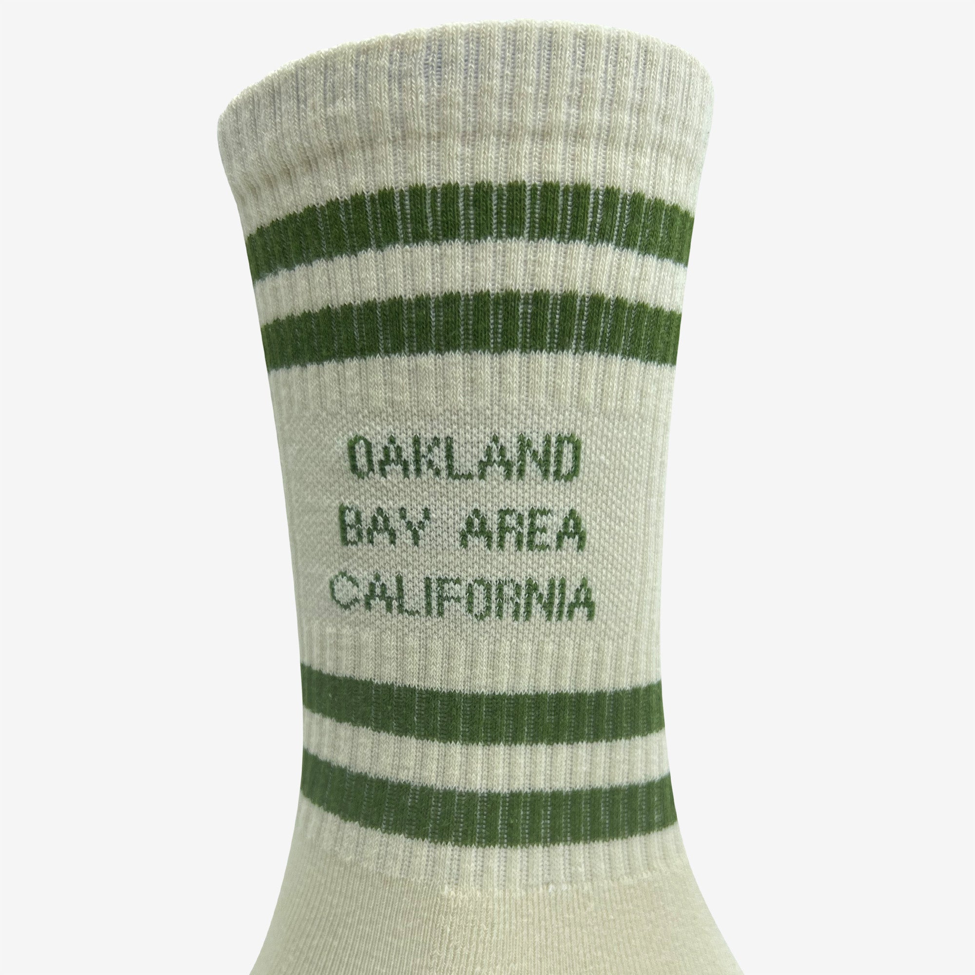 A close-up image of the Bay Area Stripes Sock by Oaklandish, featuring green stripes at the top. Between the stripes, "OAKLAND BAY AREA CALIFORNIA" is woven in green capital letters. Made from cotton or a similar fabric, this design is available in adult sizes. The plain white background highlights the sock's details.