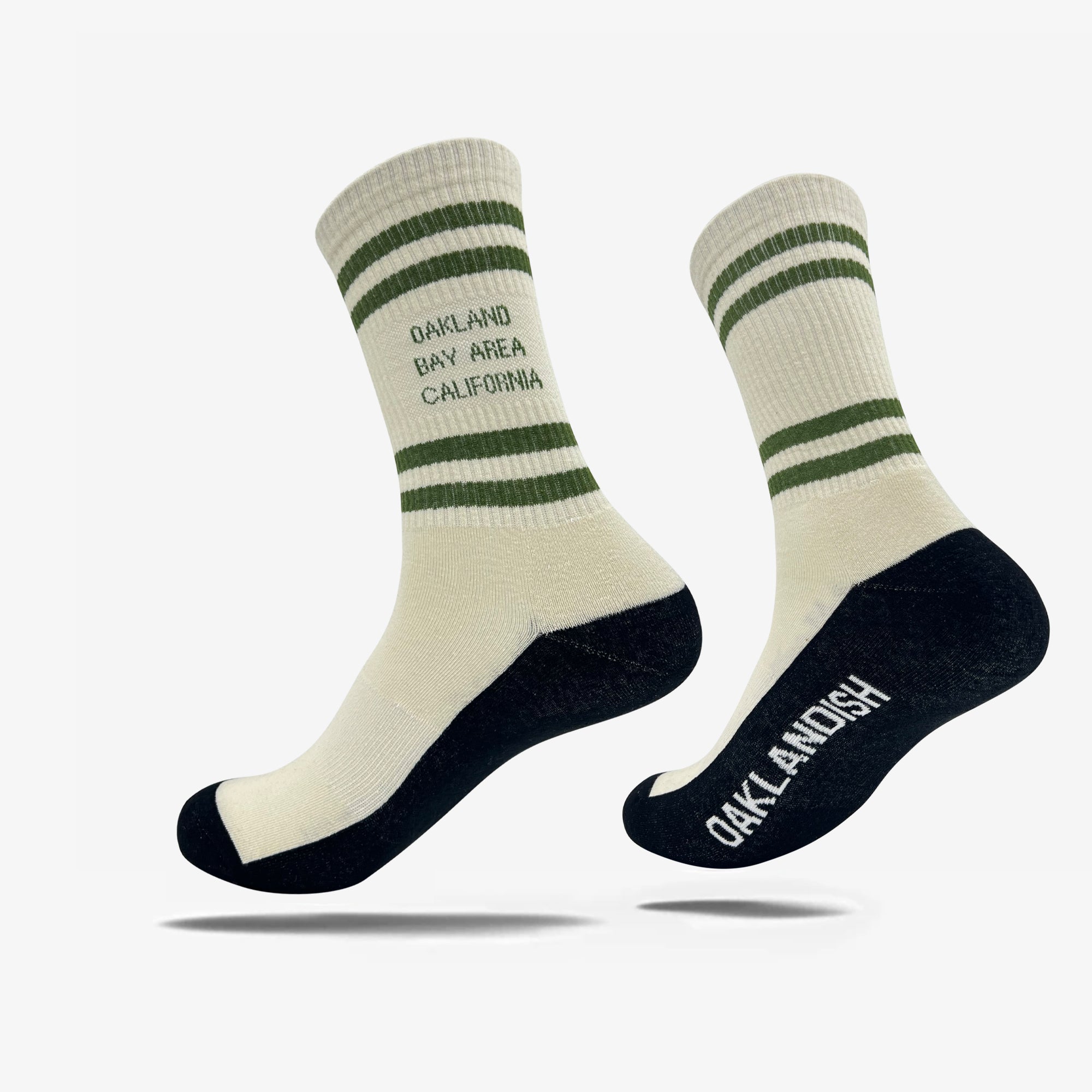 Introducing the Bay Area Stripes Sock by Oaklandish: a pair of white cotton crew socks adorned with green stripes at the top and the words "OAKLAND BAY AREA CALIFORNIA" written below. These socks feature black heels, toes, and soles, with "OAKLANDISH" printed in white on the soles. Perfect for adults, these Oaklandish logo crew socks add flair to any outfit.
