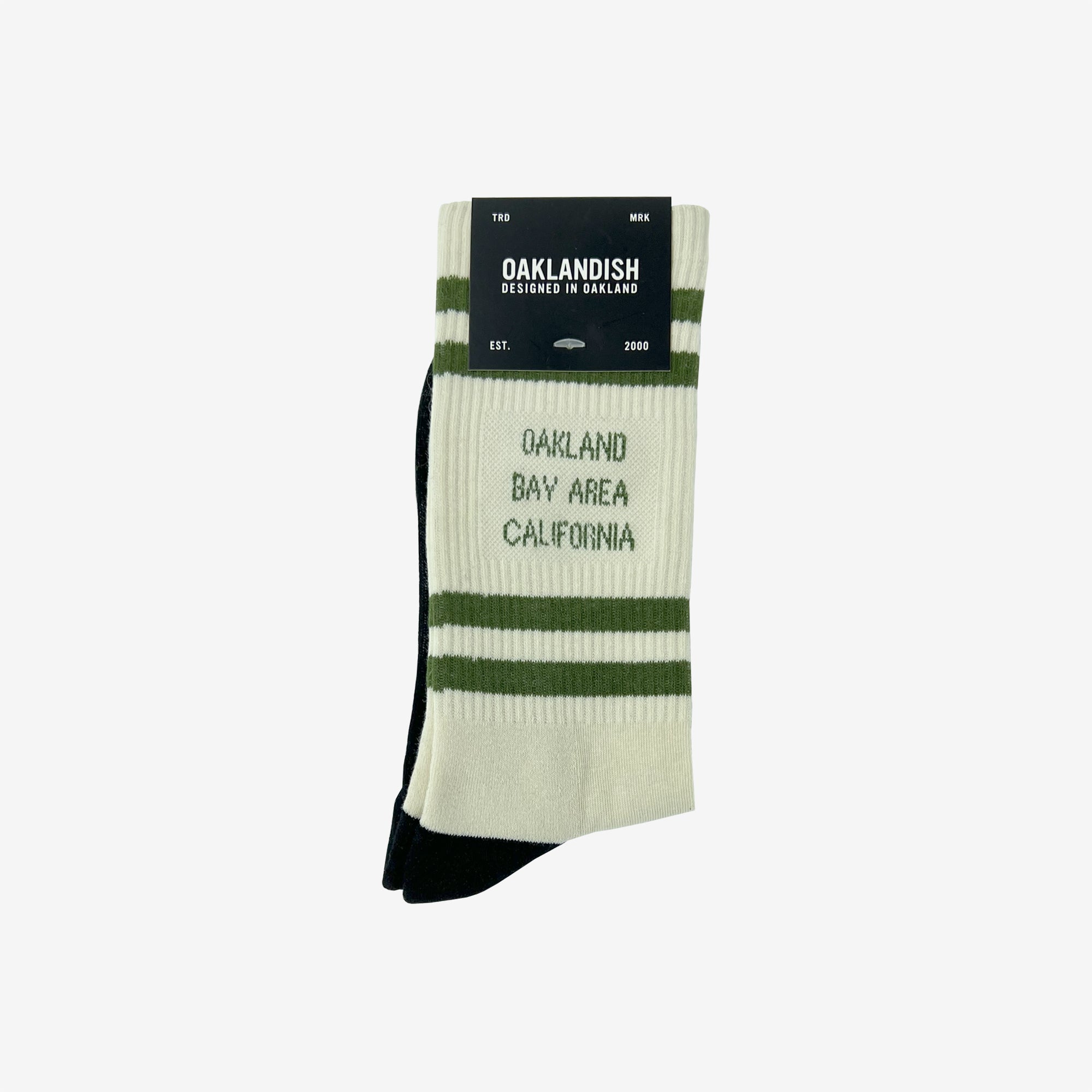 A pair of Oaklandish Bay Area Stripes Socks, featuring white crew socks with green stripes at the top, is displayed against a plain background. The adult-sized socks showcase the text "OAKLAND BAY AREA CALIFORNIA" in green. Attached to the cotton socks is a black and white cardboard tag labeled "OAKLANDISH" with additional branding information.
