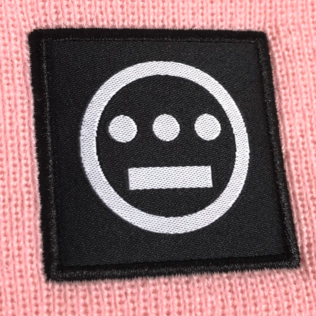 A pink knit beanie, called the Hiero Logo Cuff Beanie from Hieroglyphics, is adorned with a black square patch featuring a white geometric design. The design includes a circle containing three dots in a row above a horizontal line, representing the hip-hop collective's logo.
