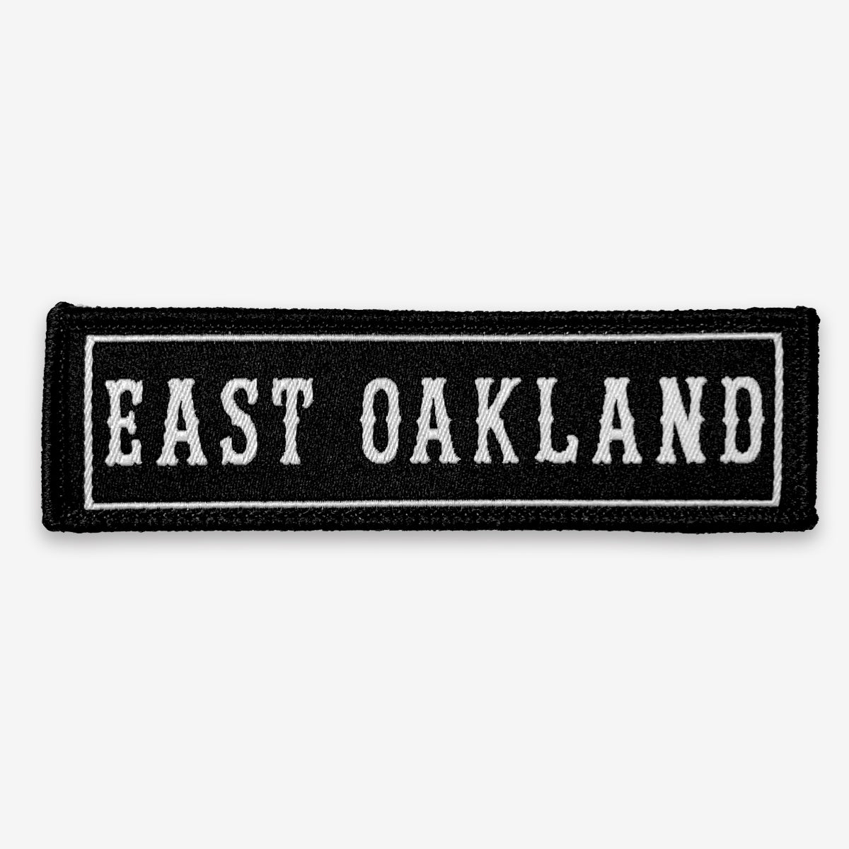 Black rectangle fabric iron patch, outlined with white line and capitalized word mark  EAST OAKLAND.”