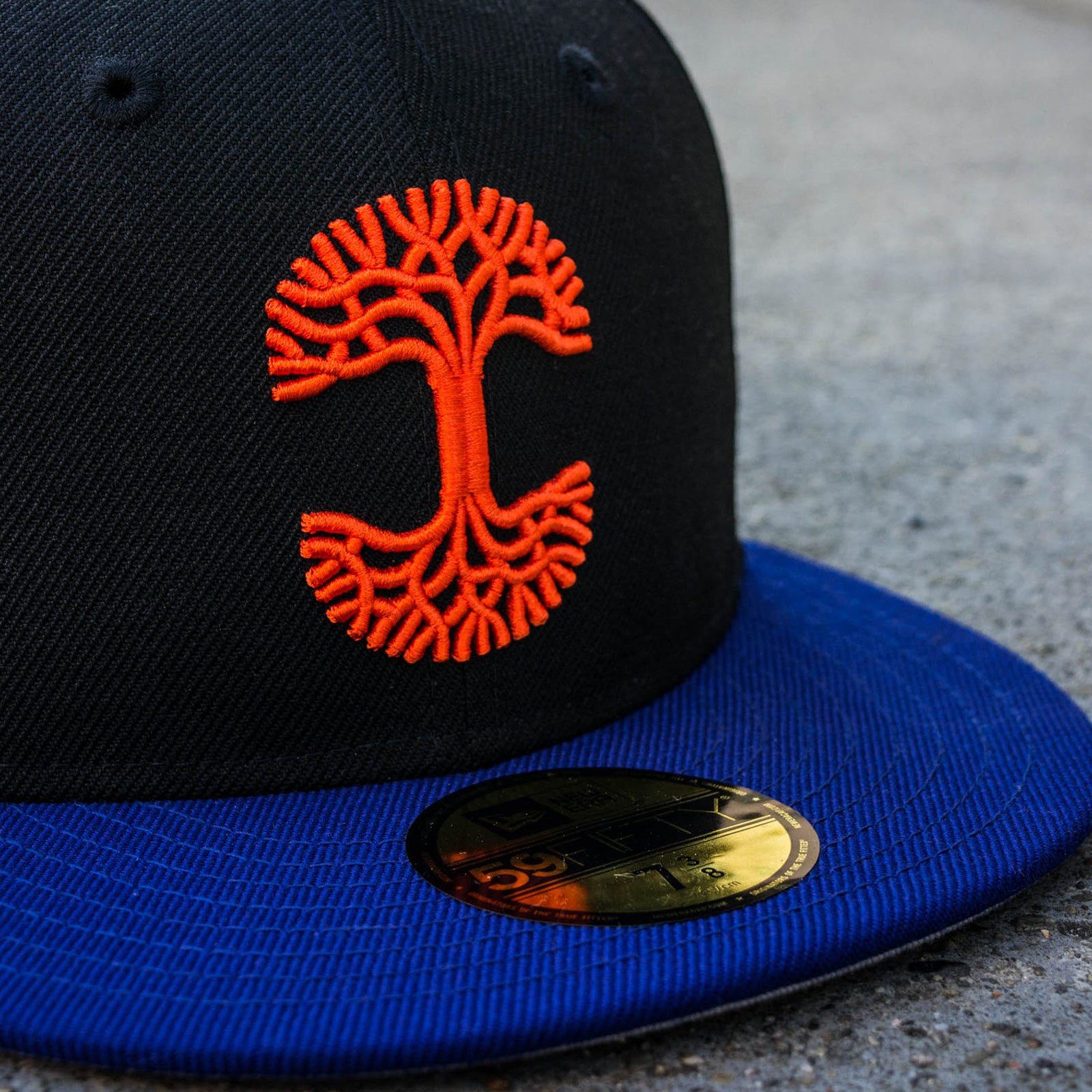 Close-up red embroidered Oaklandish logo on black fitted cap with New Era sticker on a blue bill.