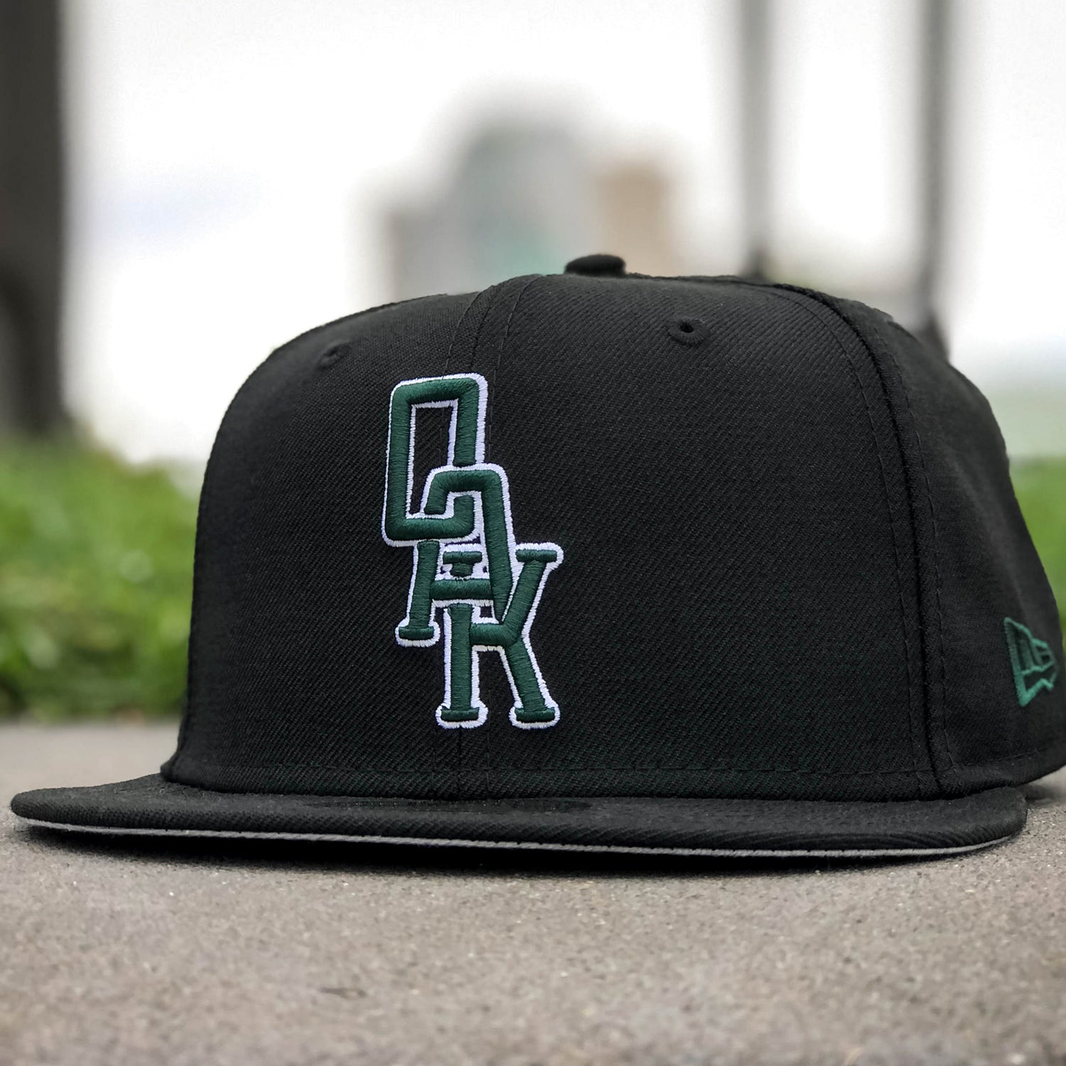A black New Era OAK 59FIFTY Fitted Cap by Oaklandish, showcasing an elevated green and white embroidered "OAK" logo on the front, is displayed on a concrete surface with a grassy area and blurred buildings in the background, adding depth to the image.