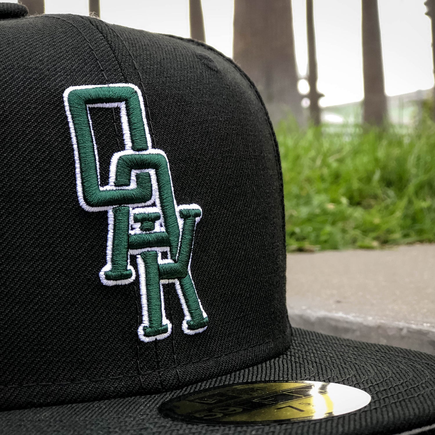 The New Era OAK 59FIFTY Fitted Cap by Oaklandish showcases a green and white embroidered "OAK" logo on the front, with the letters intertwined. A metallic gold sticker adorns the brim. The background features a blurred outdoor setting with greenery and partially visible large poles.