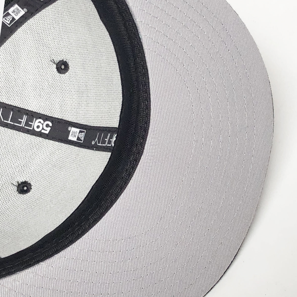 Close-up view of the inside of a New Era OAK 59FIFTY Fitted Cap by Oaklandish, revealing the grey fabric and black sweatband with branding details. The cap includes black grommets and size stickers, along with an embroidered OAK logo and intricate stitching visible on the hat’s inner surface.