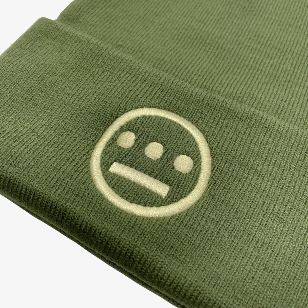 Close-up of the Hiero Classic Cuff Beanie by Hieroglyphics in olive green, featuring a simple, light beige embroidered logo. The design, reminiscent of hieroglyphics, includes a slightly rounded horizontal line beneath three small vertical rectangles enclosed within a circle, resembling a minimalist face. The background is plain white.