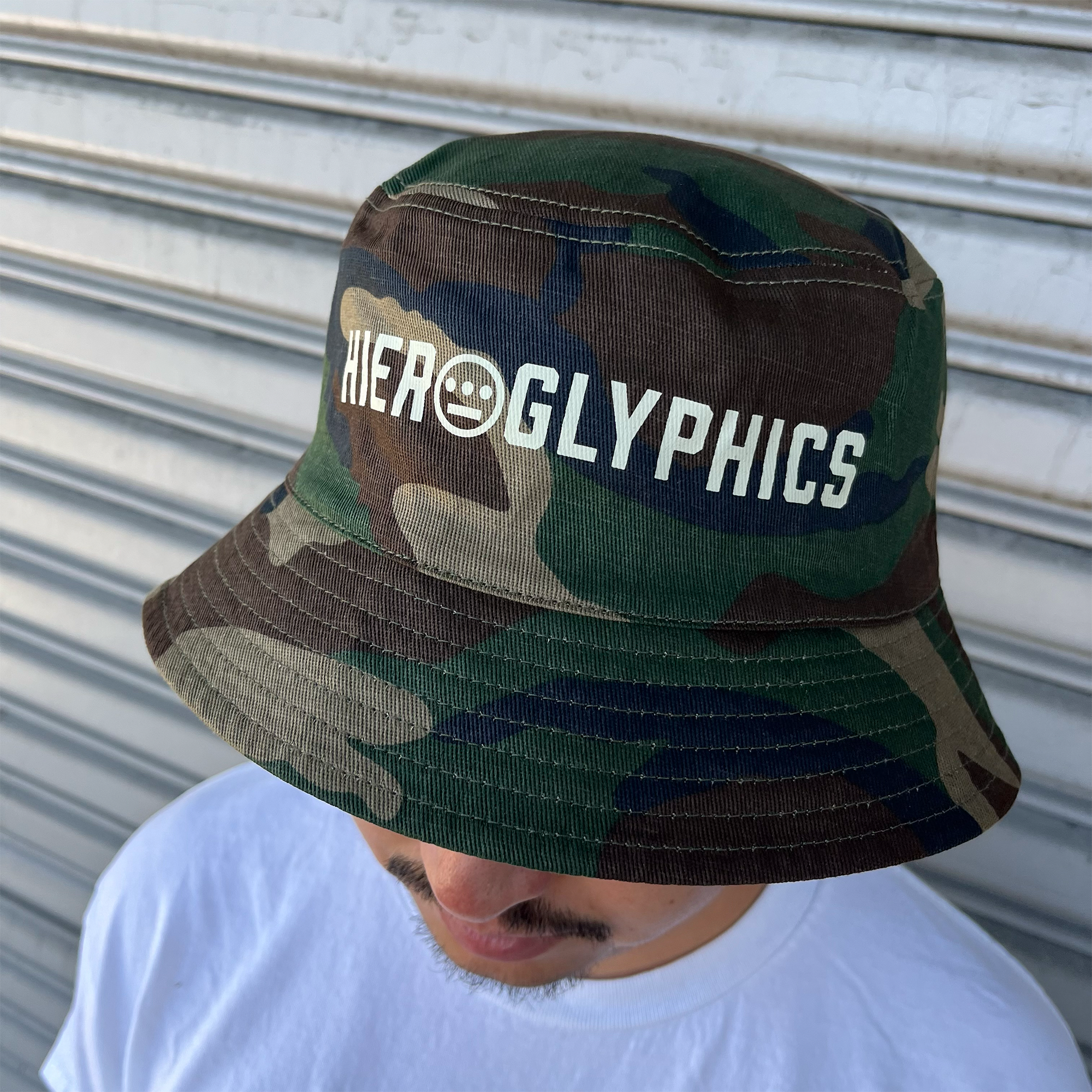Man standing outdoors, wearing a Woodland Camo bucket hat with a Hieroglyphics Hip Hop Crew wordmark on the front crown.