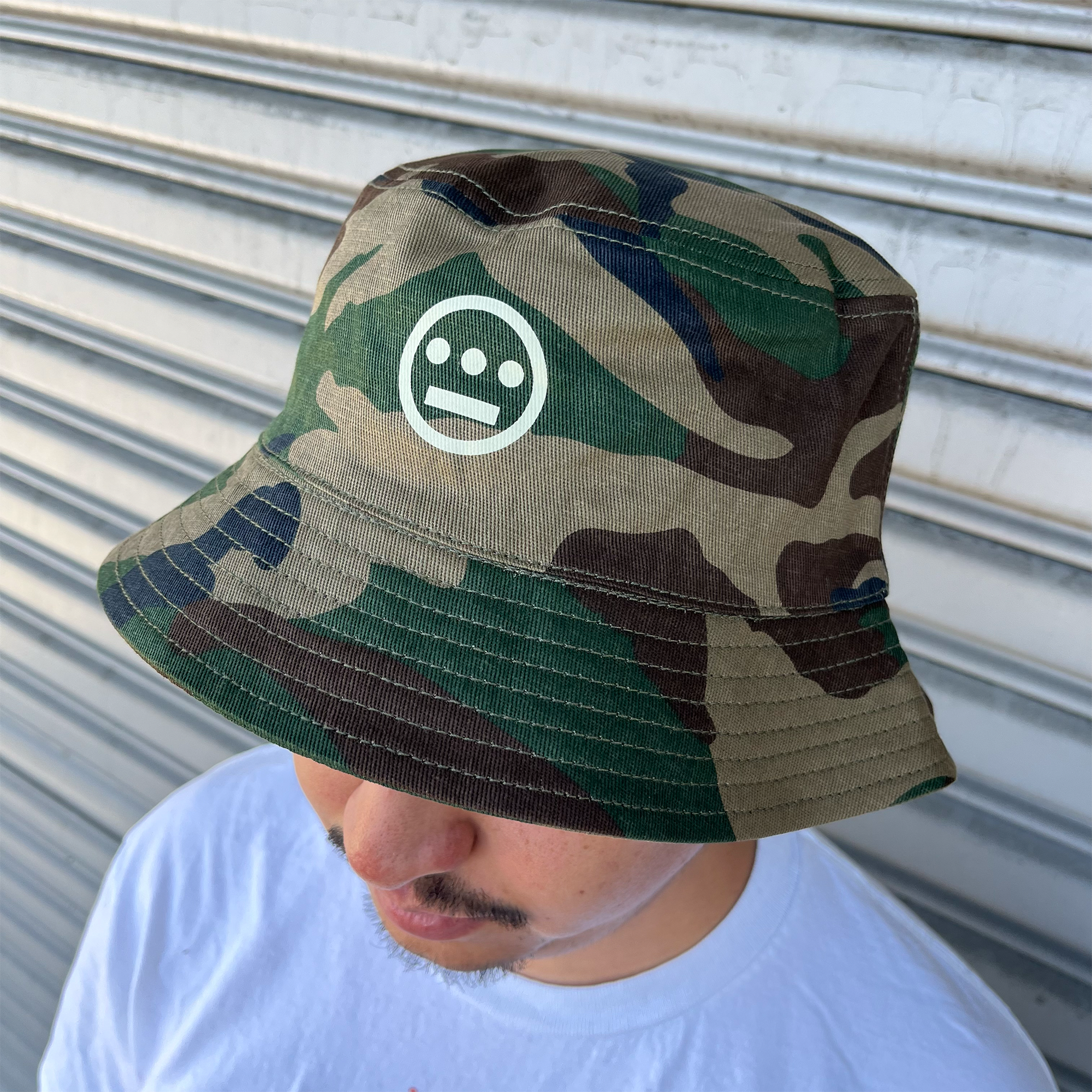 Man standing outdoors, wearing a Woodland Camo bucket hat with a Hieroglyphics Hip Hop Crew logo on the front crown.