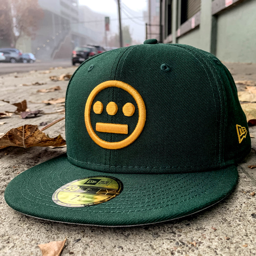 Green New Era cap with gold embroidered Hieroglyphics hip-hop logo on the crown on an outdoor sidewalk.