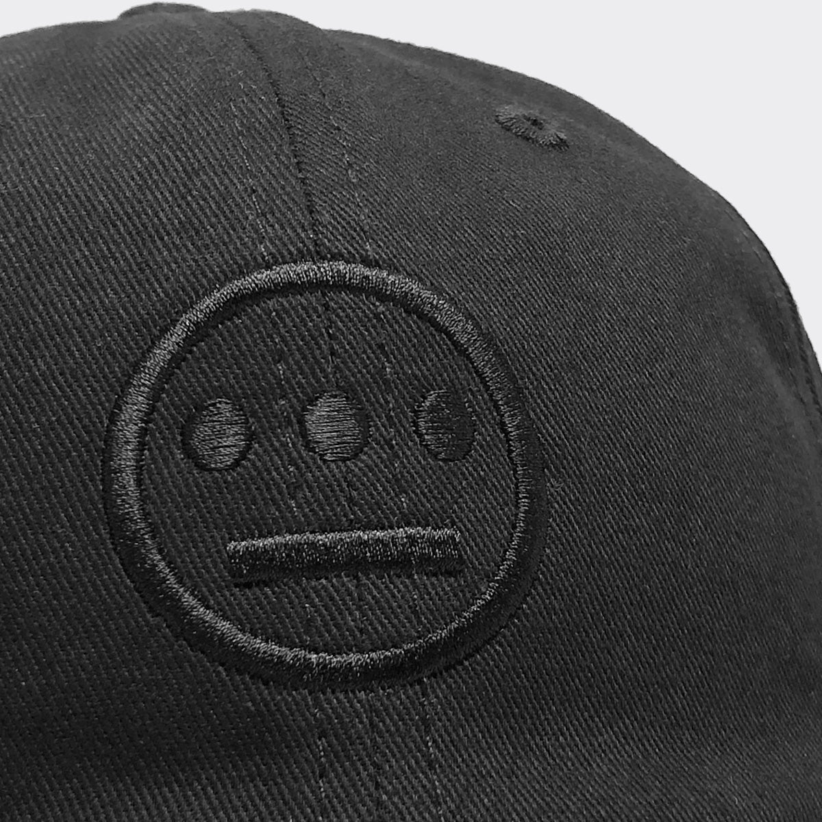 Close-up of the Hiero Logo Dad Hat by Hieroglyphics, showcasing an embroidered logo in matching black thread. The minimalist face design, reminiscent of hieroglyphics, features three dots for eyes and a straight horizontal line for a mouth. It includes an adjustable strapback, all set against a plain light grey background.