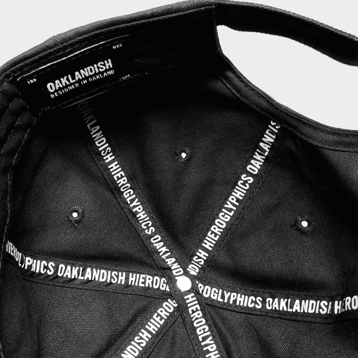 Close-up view of the inside of the Hiero Logo Dad Hat by Hieroglyphics, featuring intersecting black bands with white text. The text across the bands reads "OAKLANDISH HIERGLYPHICS." The inner label at the crown displays "OAKLANDISH DESIGNED IN OAKLAND." The cap, which has an adjustable strapback, is detailed with small ventilation holes.