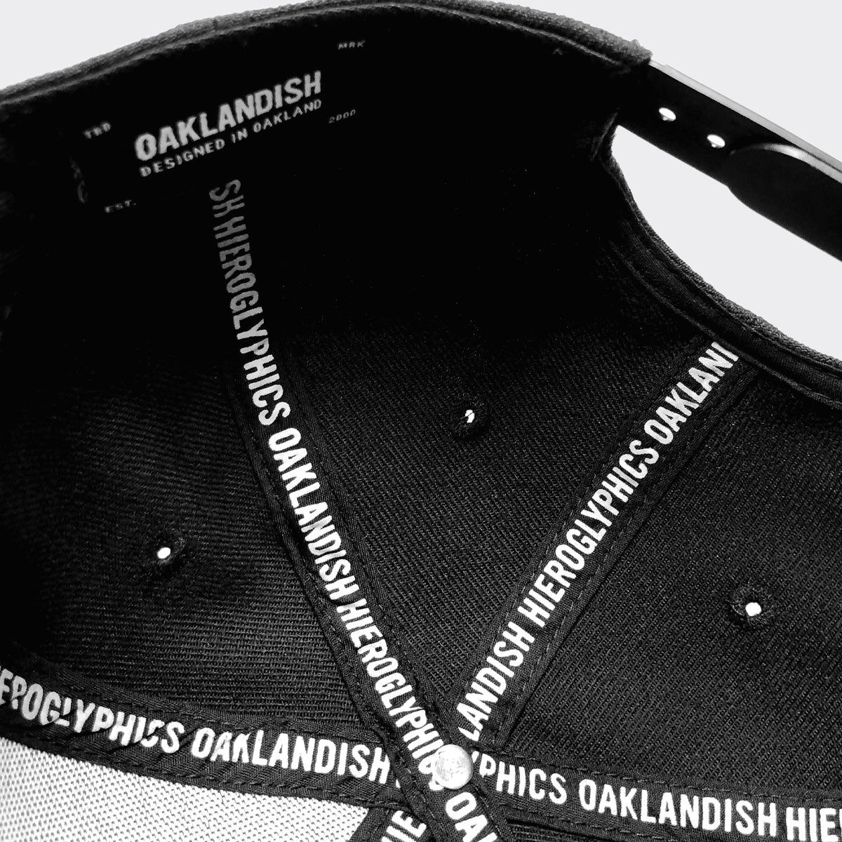 Close-up image of the inside of a Hiero Logo Snapback by Hieroglyphics. The interior features white embroidered text on black fabric strips that cross the cap's structure. The text reads “HIEROGLYPHICS OAKLANDISH” repeatedly. The snapback also has a label with the same phrases and "DESIGNED IN OAKLAND.
