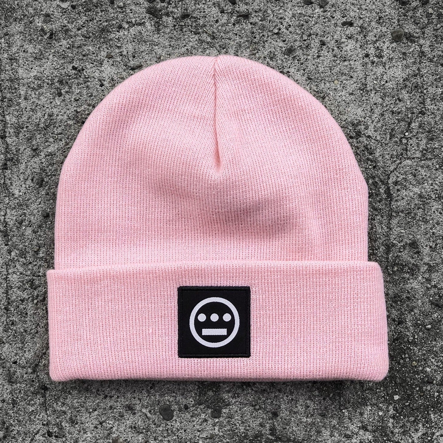 The Hieroglyphics Hiero Logo Cuff Beanie, in light pink knit, is showcased on a concrete surface. It features a folded brim with a black patch sewn onto the front. The patch displays a white circle with three horizontal dots centered above a line, adding a touch of modern hieroglyphics to your hip hop style.