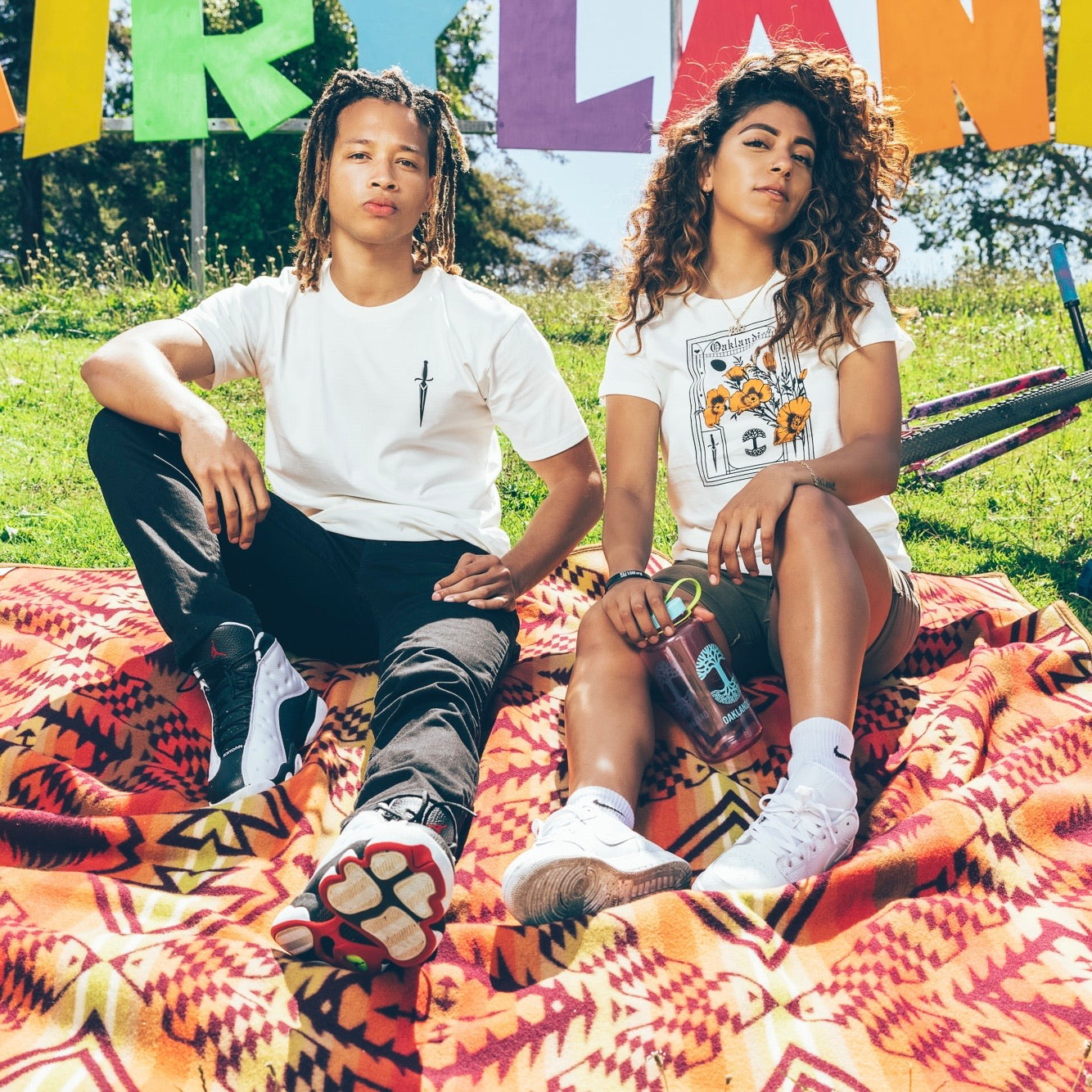 A man and woman sitting in the park on a picnic blanket wearing Oaklandish Blossom t-shirts.