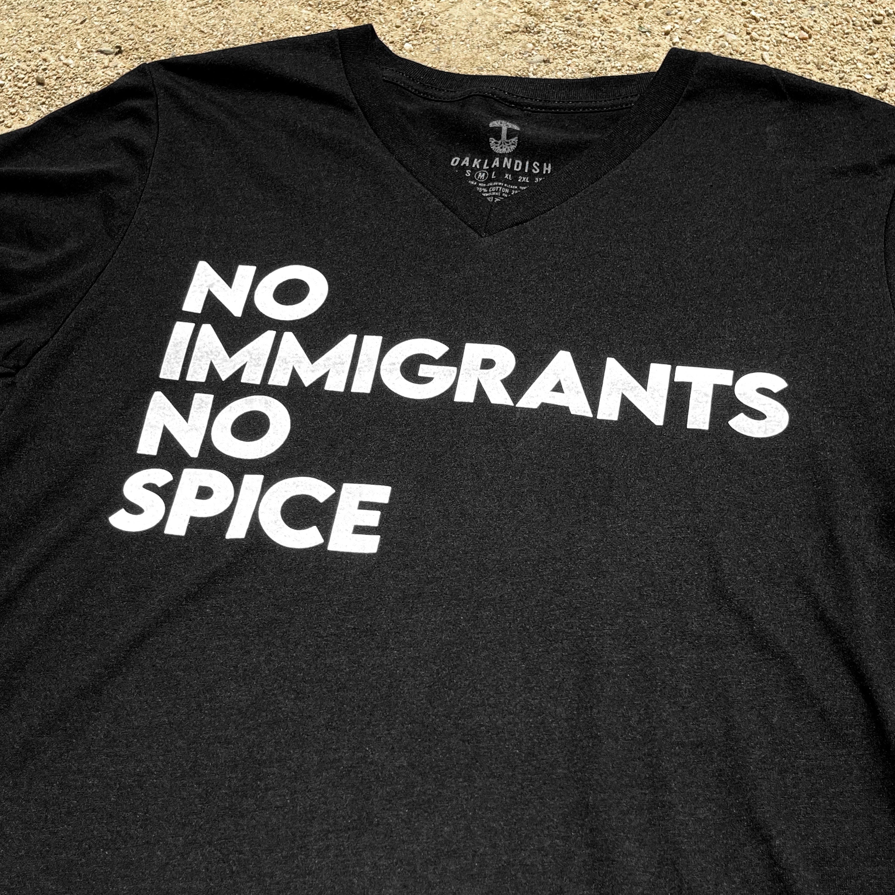 Close-up of black V-neck t-shirt with white No Immigrants, No Spice wordmark laying on sand.