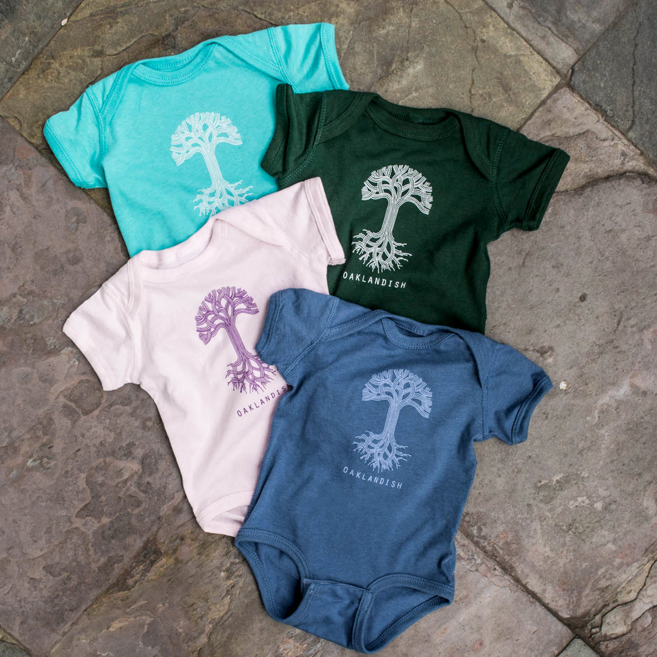 Four "Infant Oaklandish Classic Logo One-Piece" rompers are arranged on a stone surface. From top left, they come in light blue, dark green, pale pink, and navy blue. Each 100% cotton romper features a tree design with the text "OAKLANDISH" beneath it in corresponding colors: white on the blue and green ones, purple on the pink one, and white on the navy one.